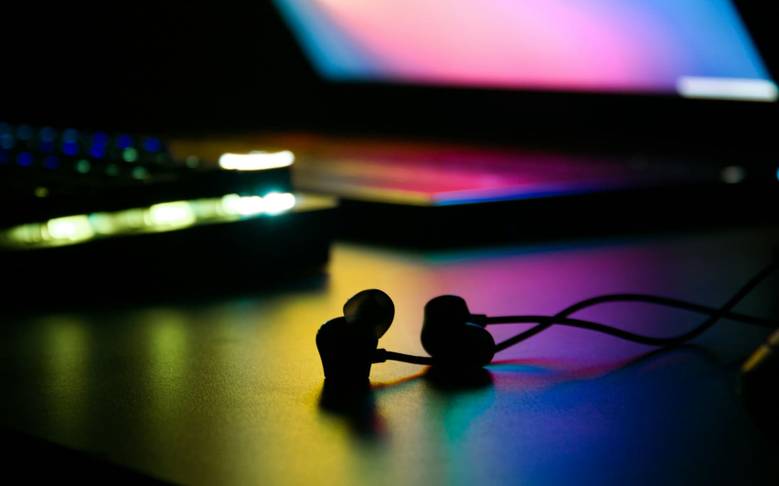 A pair of wired earbuds lies on a dark desk, lit in yellow, pink and blue by a laptop screen and a glowing backlit keyboard