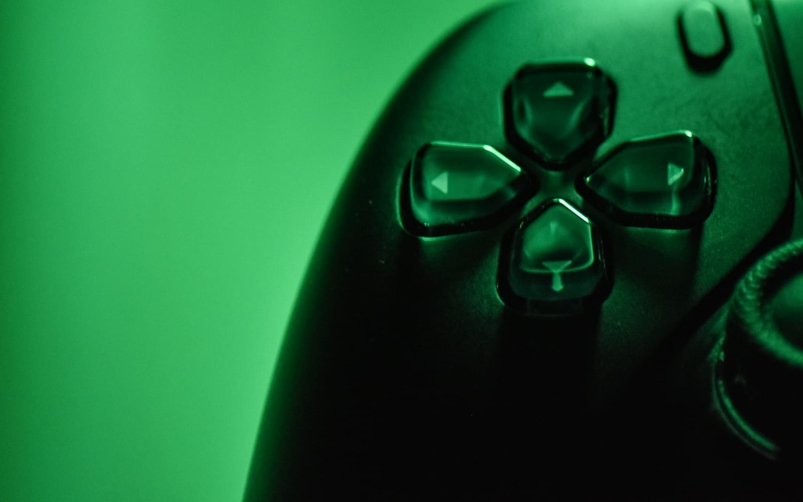 Close-up of a black game controller’s directional pad, its glossy arrow buttons lit by green light against a soft green background