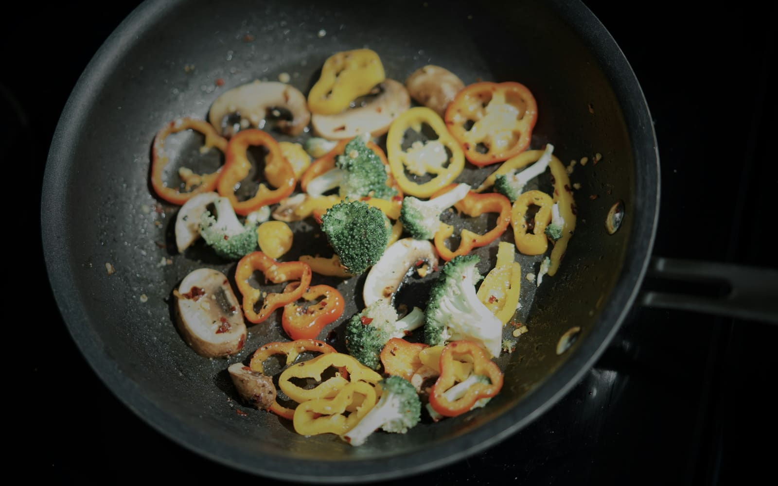 Sliced red and yellow peppers, mushrooms and broccoli florets cooking in a black nonstick frying pan on a dark stovetop, seen from above