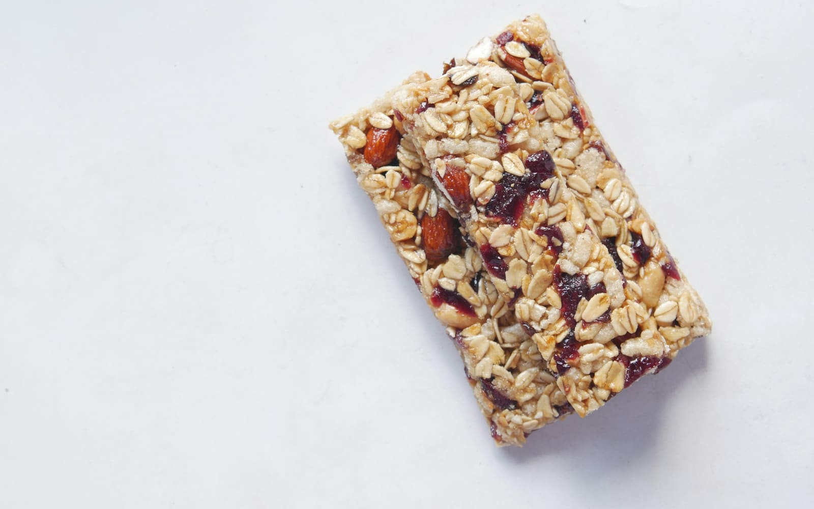An oat bar studded with almonds and dried cranberries resting on top of a second bar on a plain white surface