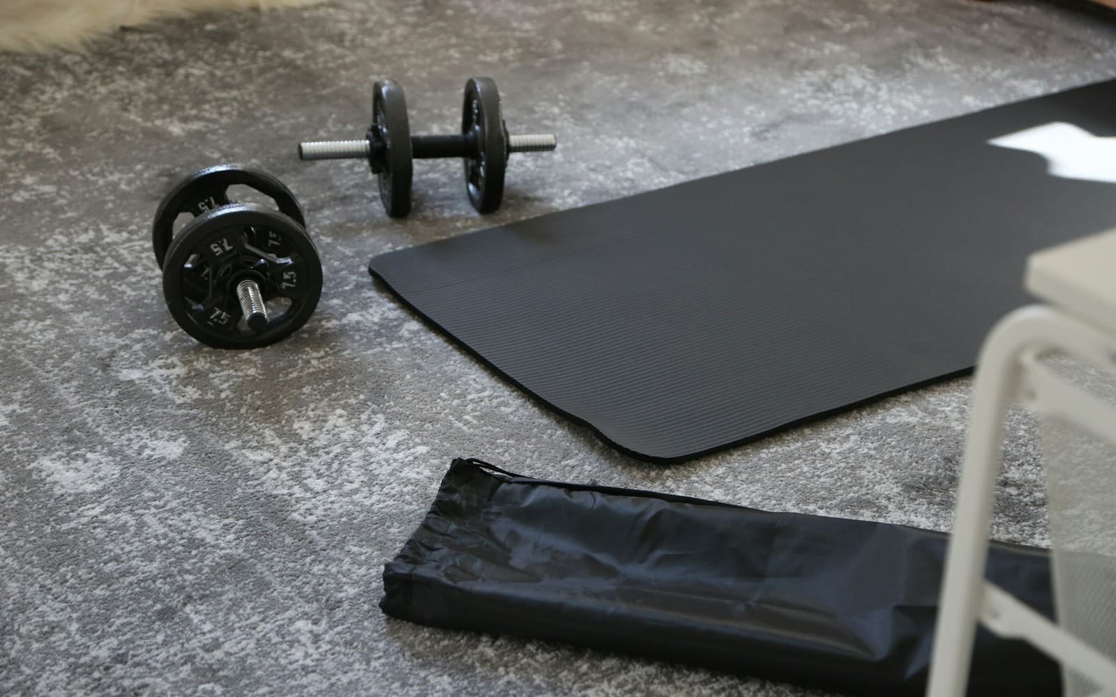 A pair of black iron dumbbells beside a black exercise mat and its carry bag on a gray rug, with a white chair leg in the corner