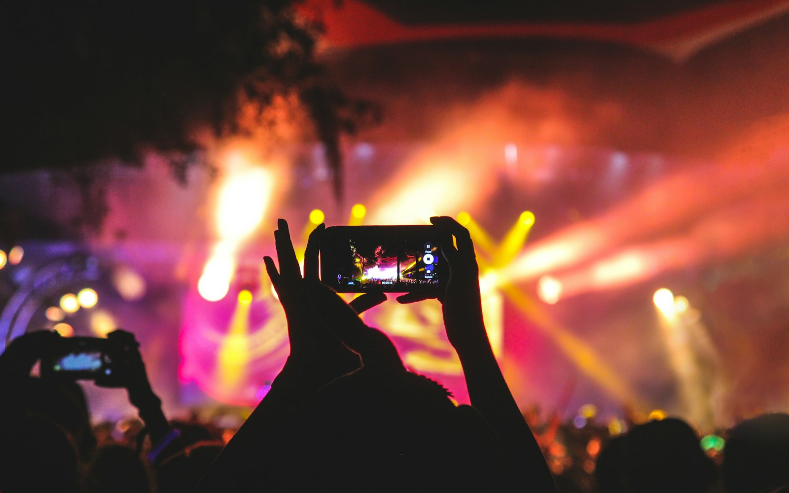 Silhouetted hands holding up a phone to film a concert stage lit by pink and yellow spotlights, with another phone raised nearby