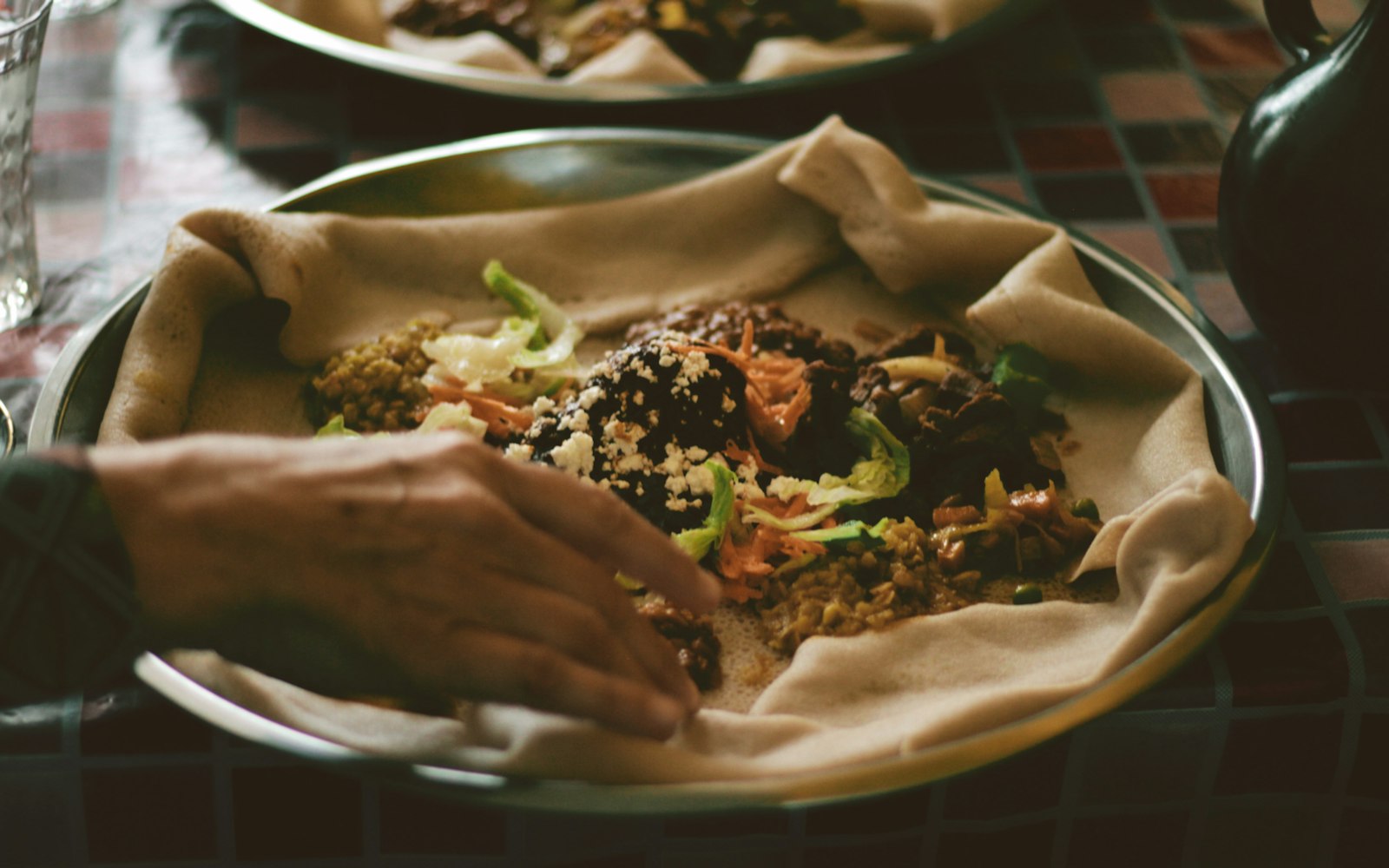 A hand reaching toward a metal platter lined with injera and topped with stews, shredded vegetables and crumbled cheese, on a tiled table