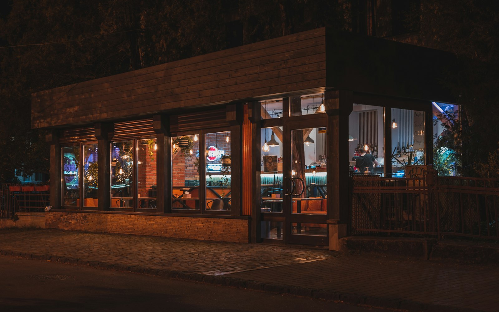 A small wood-clad restaurant at night, its large windows glowing warmly onto an empty cobblestone sidewalk, with a worker visible inside