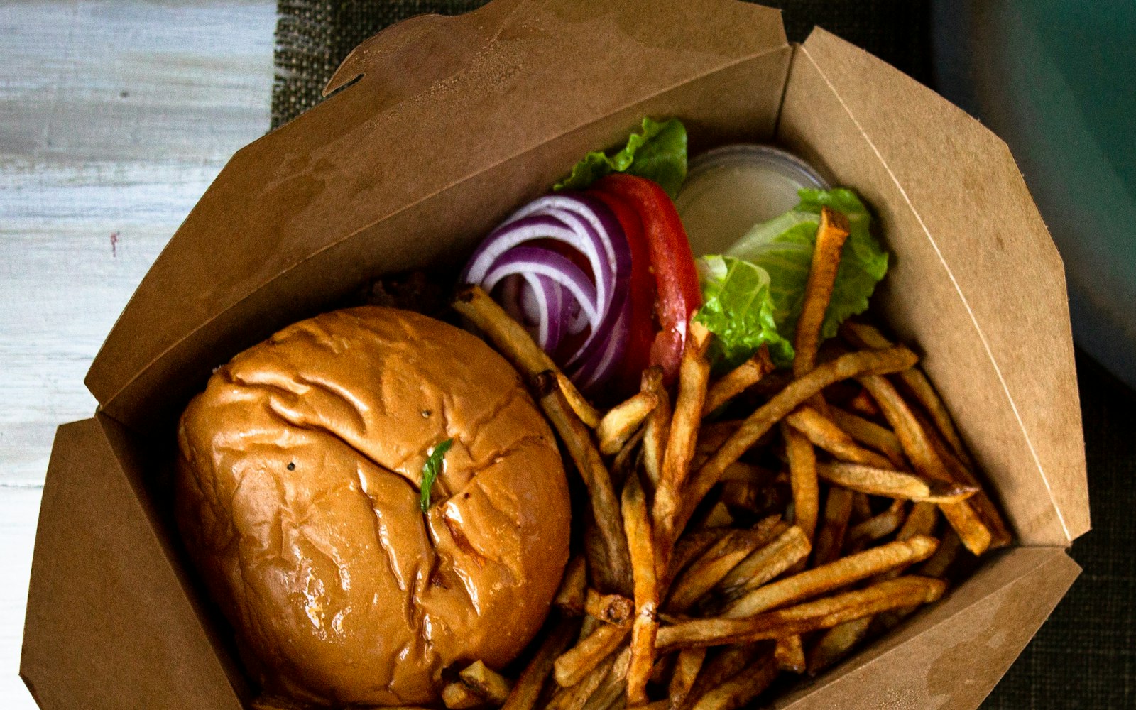Overhead view of an open kraft paper takeout box holding a burger, sliced red onion, tomato, lettuce and a heap of fries