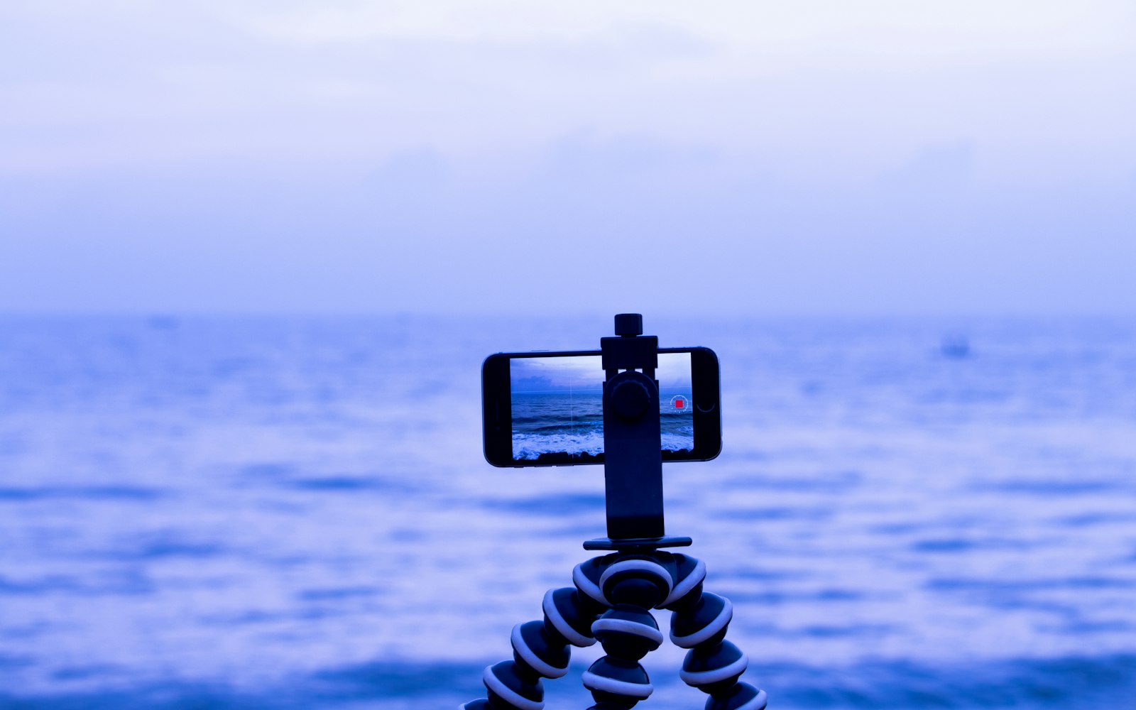 A smartphone held horizontally on a small flexible tripod records the sea, its screen showing gray waves under a hazy blue sky