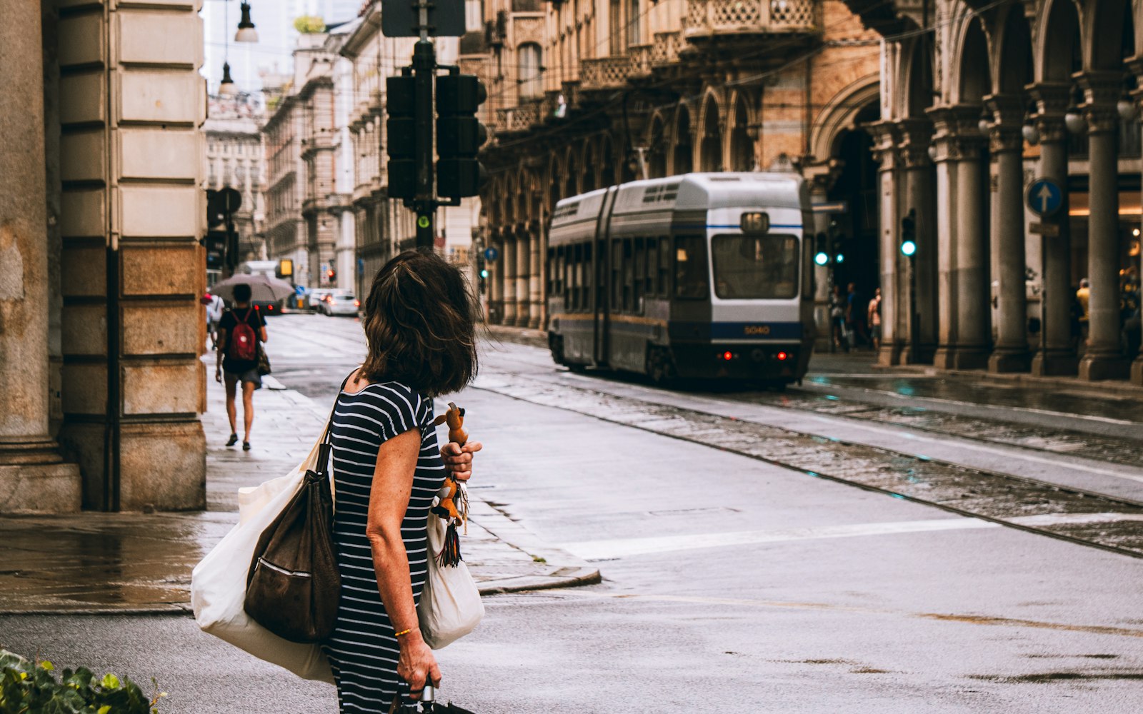 A woman in a striped dress, seen from behind with tote bags, waits at a wet street corner as a tram passes stone arcades