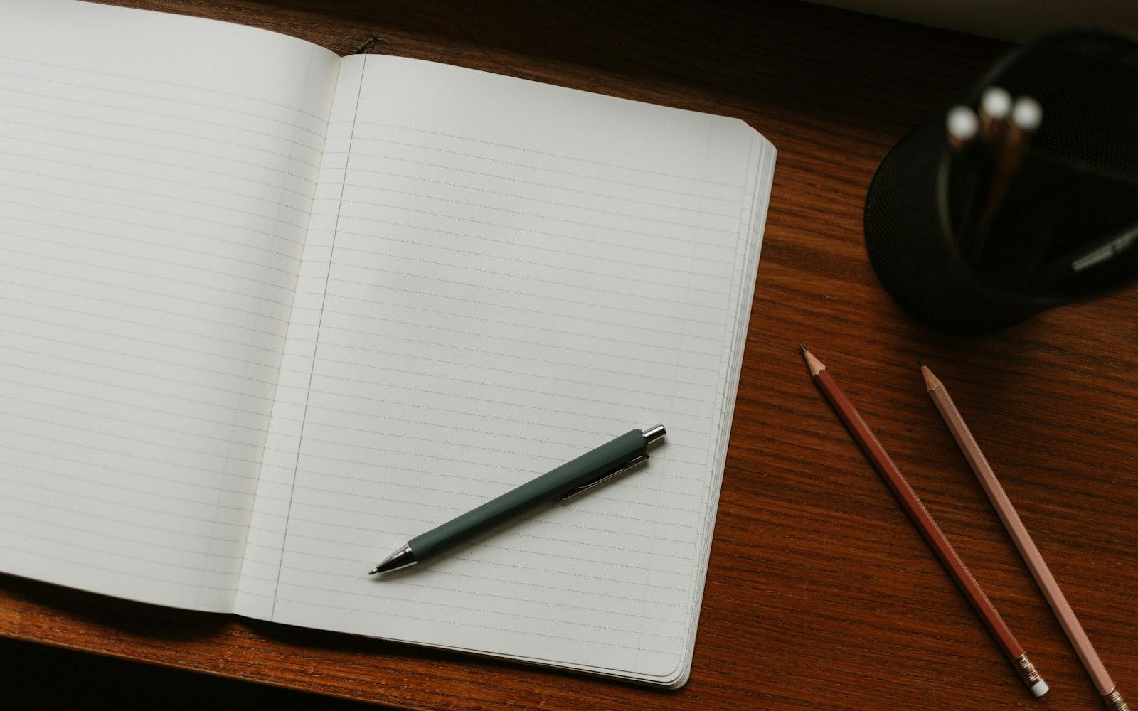 An open lined notebook with a green pen resting on it, on a wooden desk beside two sharpened pencils and a black mesh pen holder