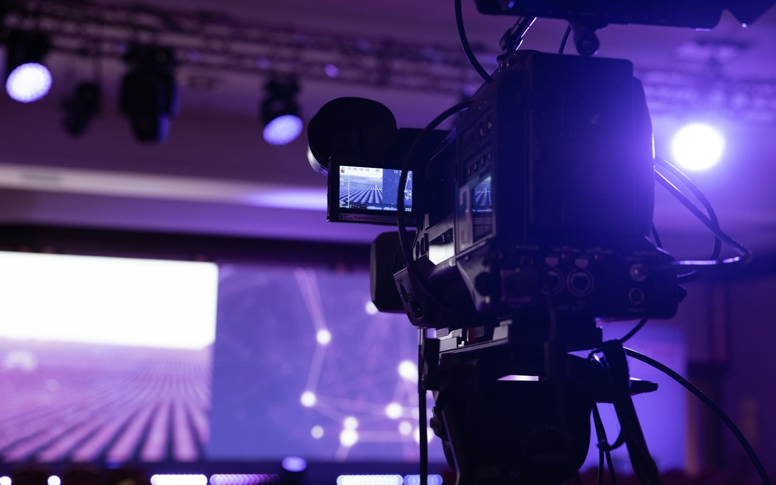 A broadcast video camera on a tripod, seen from behind, pointed at a stage lit purple by overhead spotlights and a large screen