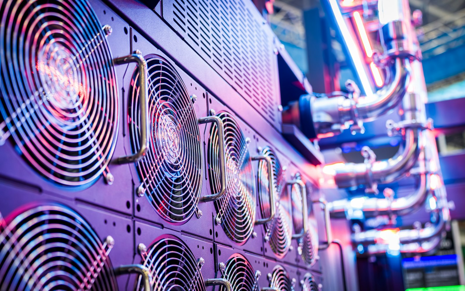 Rows of large cooling fans behind metal grilles on a server cabinet lit purple and blue, with chrome coolant pipes glowing in the background
