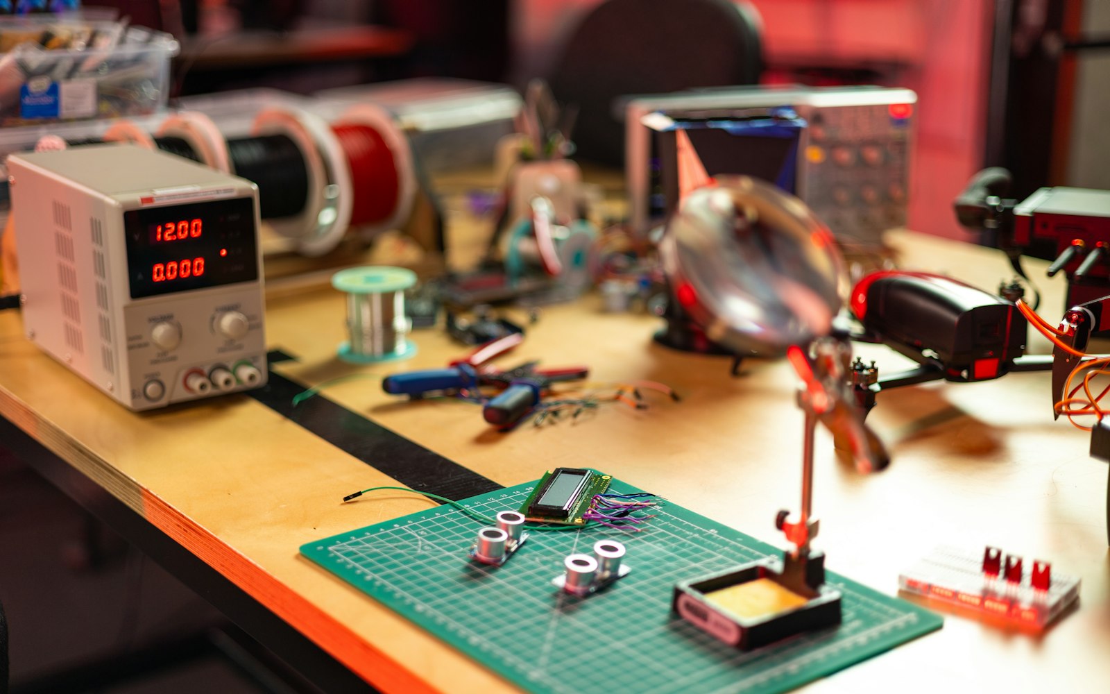 An electronics workbench with a bench power supply reading 12.00, spools of wire, pliers, solder, a magnifier and small sensors on a green cutting mat