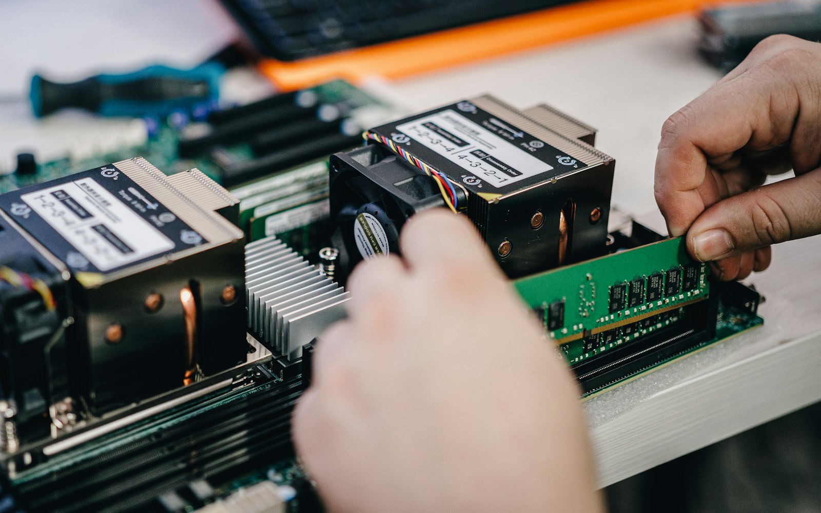 Two hands press a green memory module into a server motherboard beside two black CPU coolers on a workbench, a screwdriver blurred behind