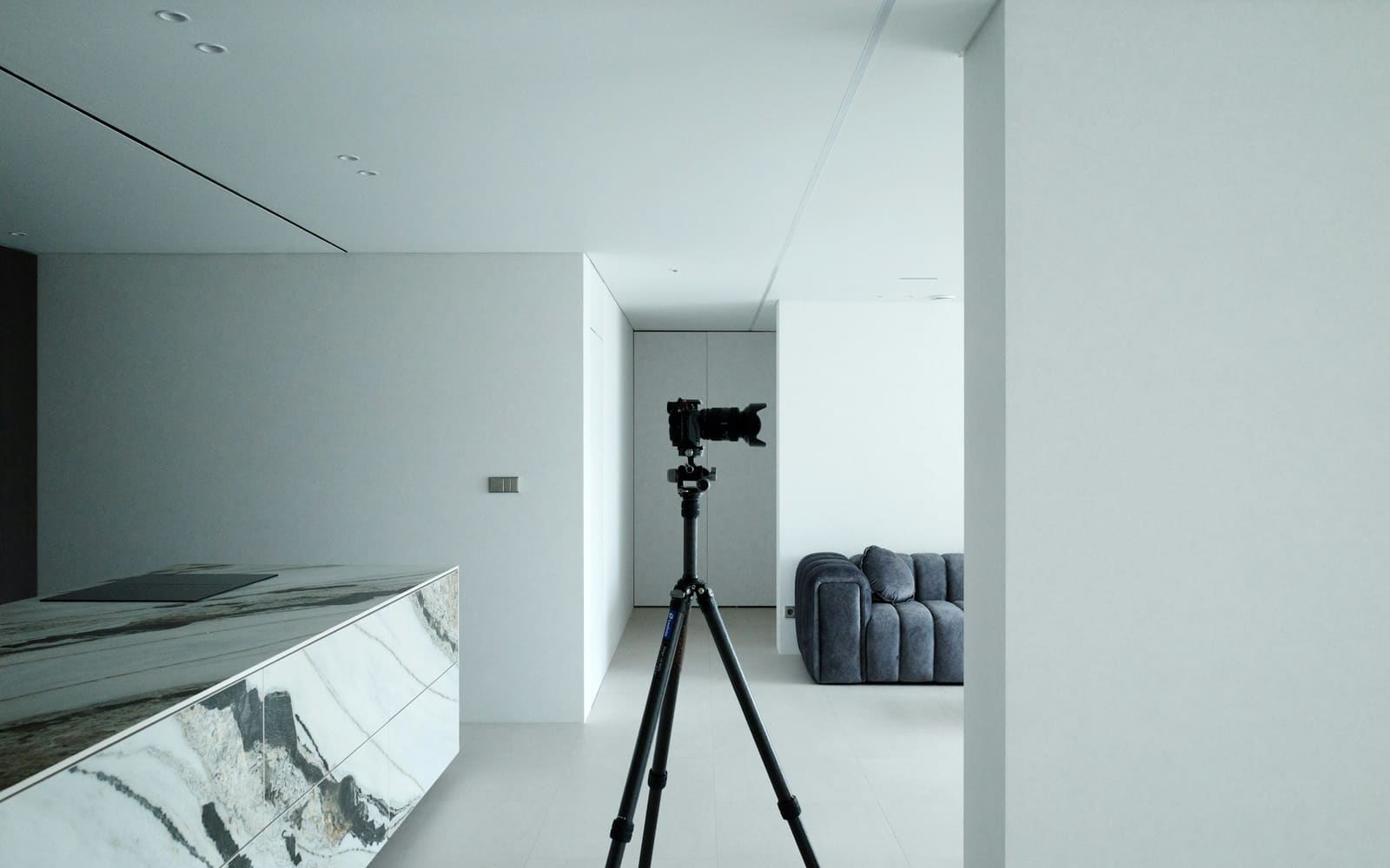 A camera on a tall tripod stands alone in a quiet white apartment, with a marble kitchen island on the left and a gray sofa in the next room