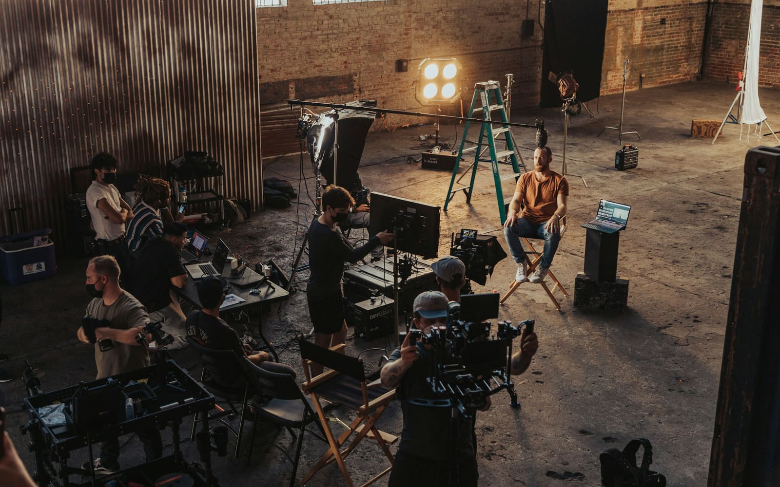 A film crew works in a brick warehouse, with camera operators, a lit lamp, a green ladder and a man seated on a director’s chair being filmed