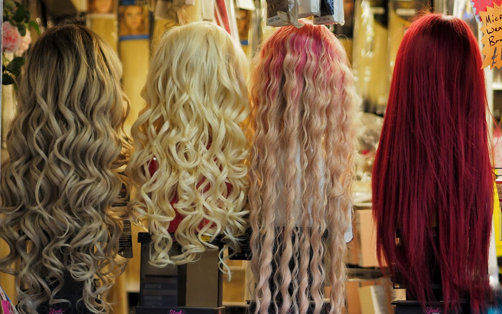 Four long wigs in ash blonde, pale blonde, pink-streaked and deep red shades sit on stands in a shop, seen from behind