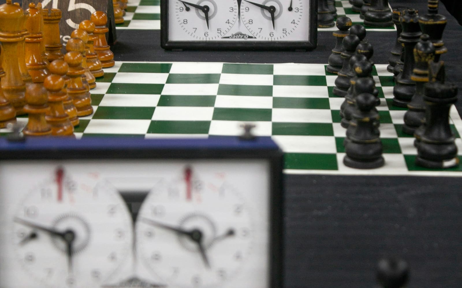 Two chess clocks stand at the ends of green and white vinyl boards, with wooden pieces lined up for games on a long table