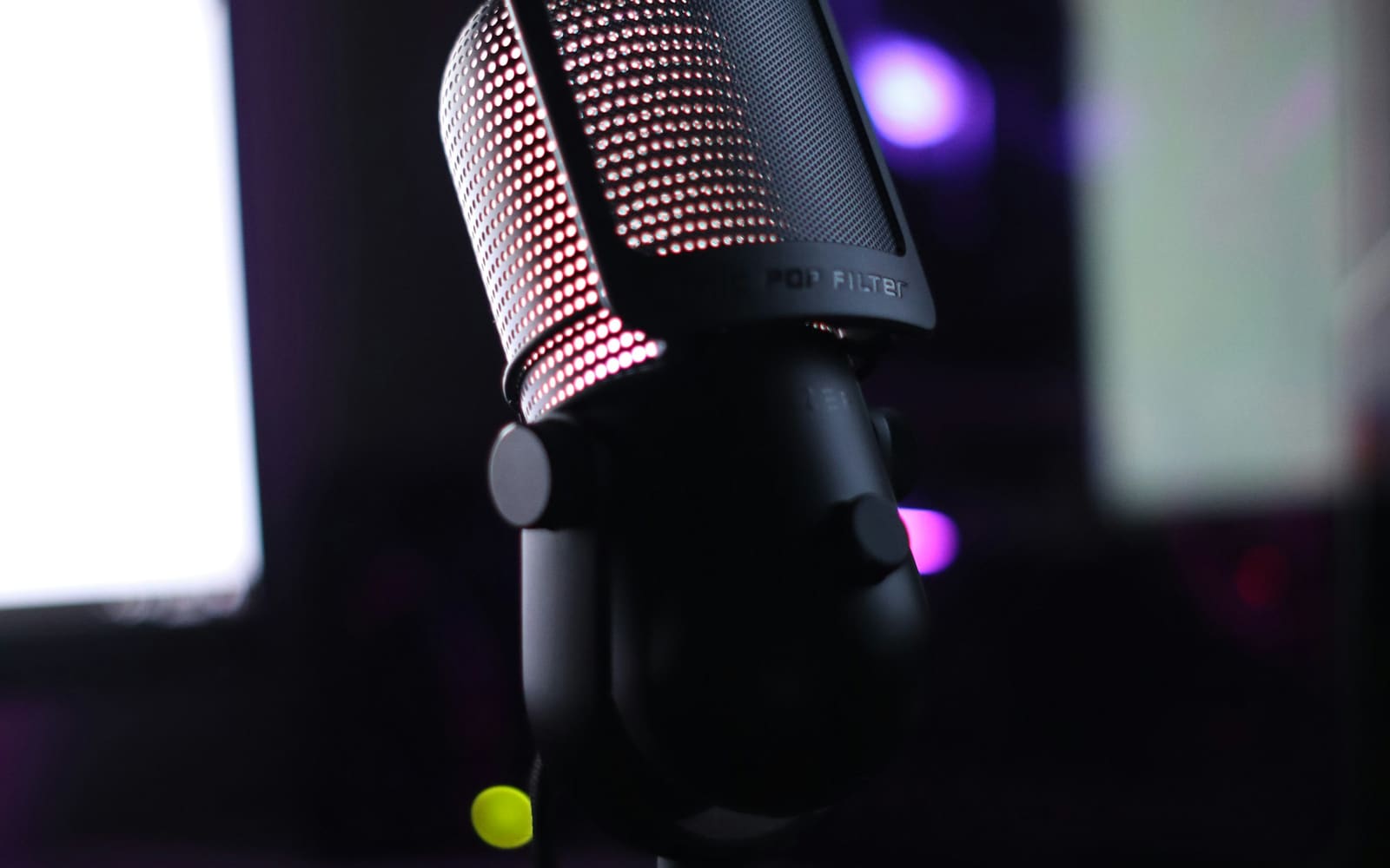 A black desktop microphone with a perforated metal pop filter glows pink from inside, in a dark room lit by blurred screens and purple light