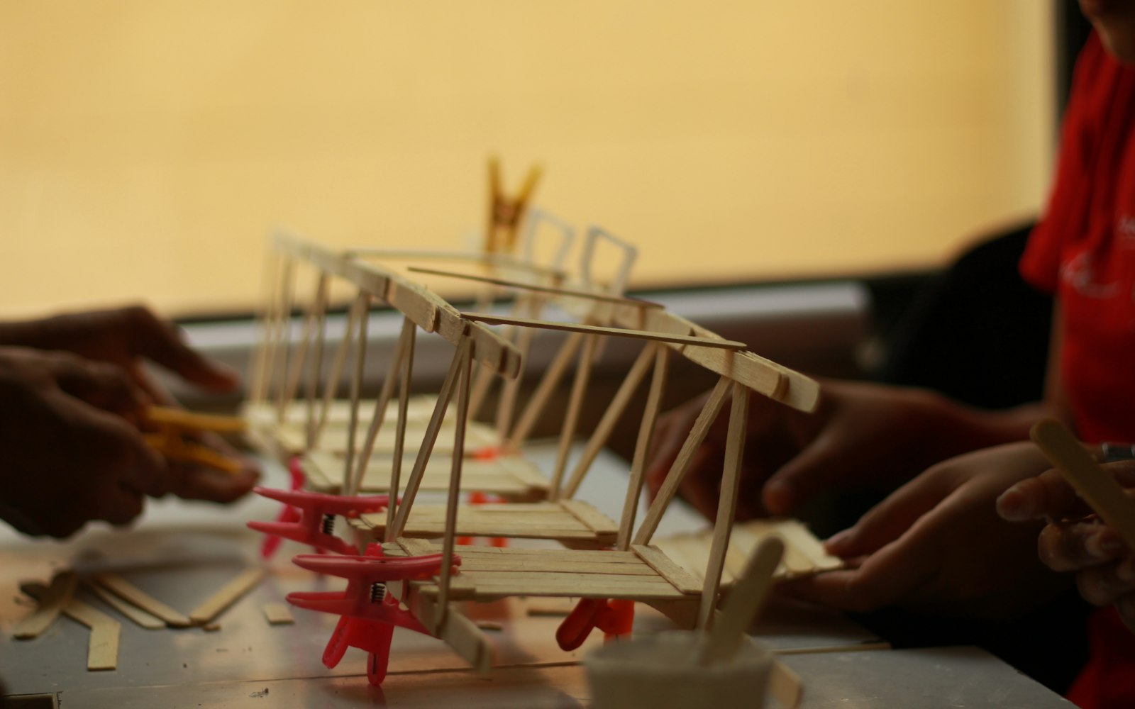 Several pairs of hands building a popsicle-stick truss model held with pink clips, with loose sticks and a glue pot on the table