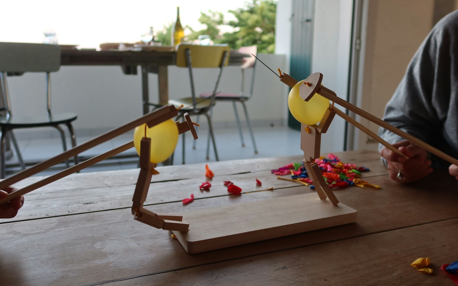 Two homemade wooden figures holding pins, each with a yellow balloon, worked by long sticks on a table scattered with popped balloons