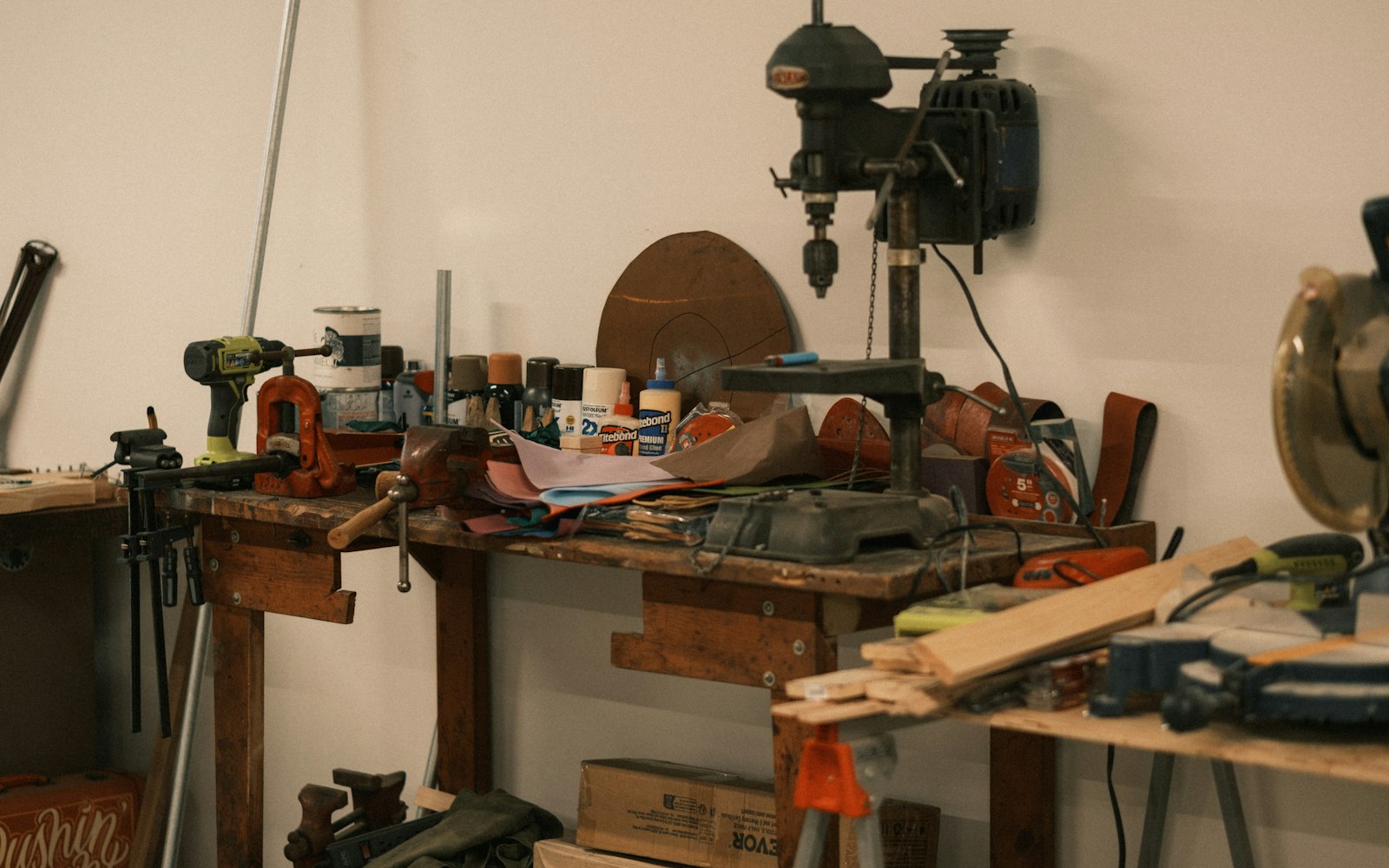 A cluttered wooden workbench in a workshop with a drill press, bench vise, cordless drill and glue bottles, and a miter saw beside it