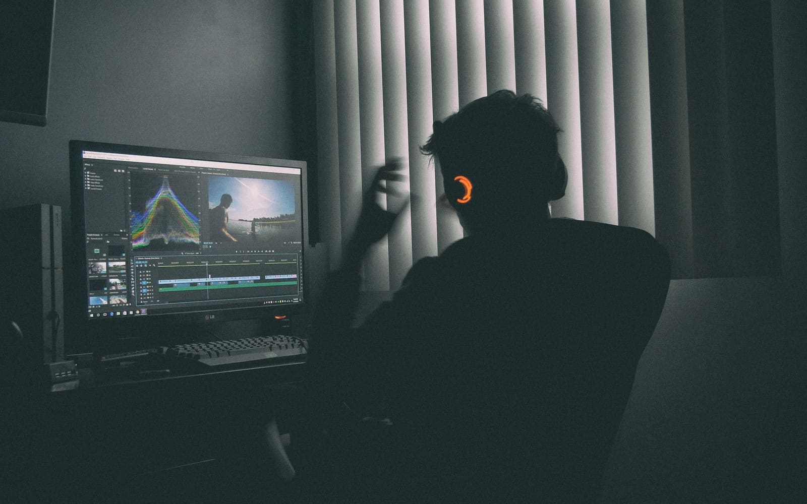 A person seen from behind in a dark room wears headphones and edits video on a desktop monitor beside window blinds