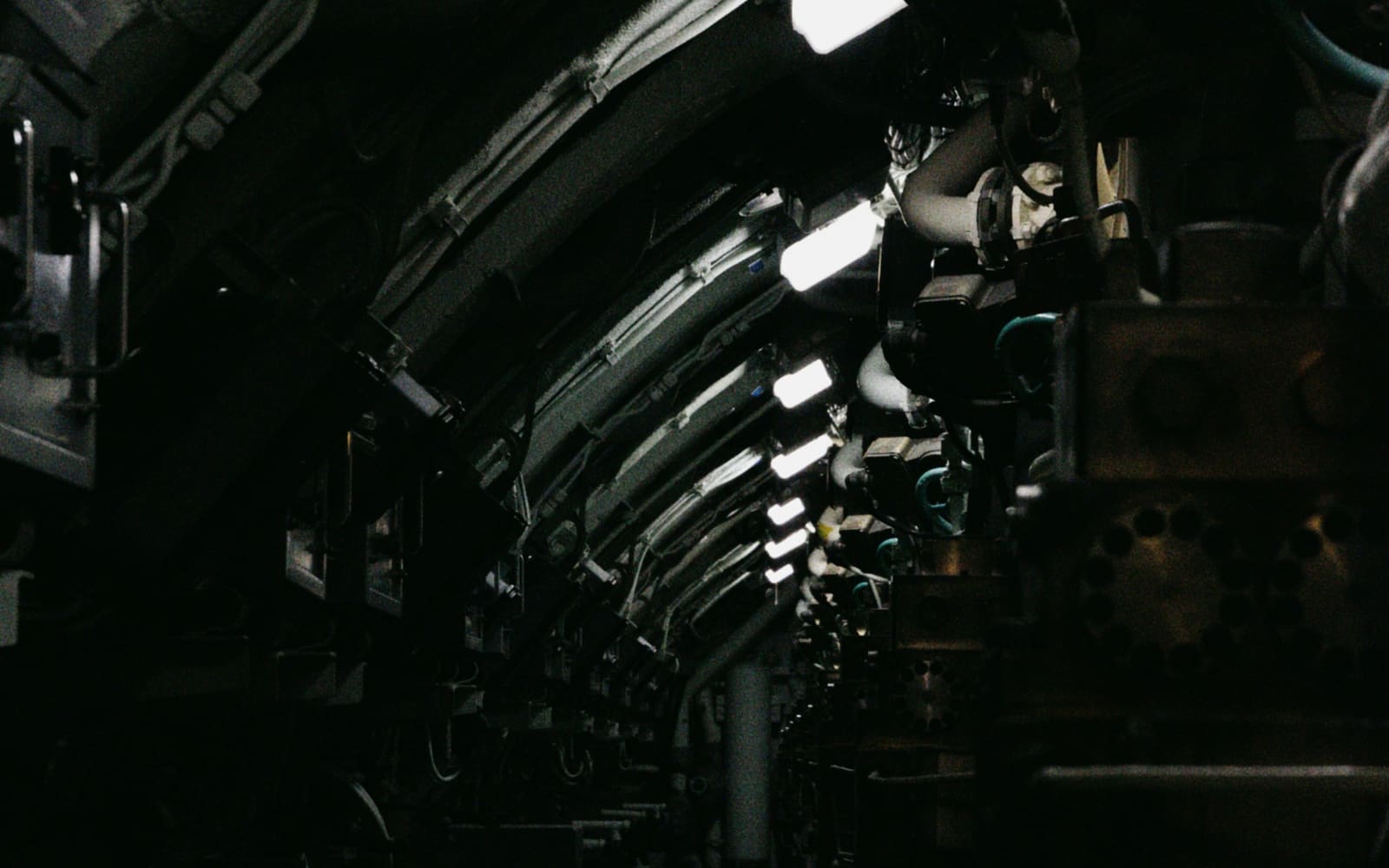 A narrow, dark submarine corridor curves away under a row of bright ceiling lamps, lined with pipes, valves and machinery