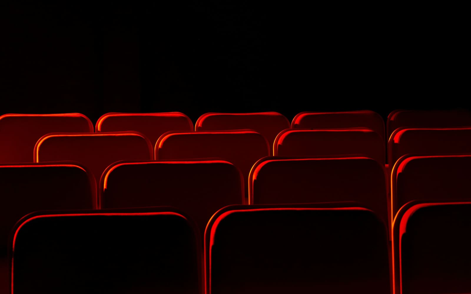 Rows of empty red cinema seats in a dark auditorium, their curved backs edged with red light against a black background