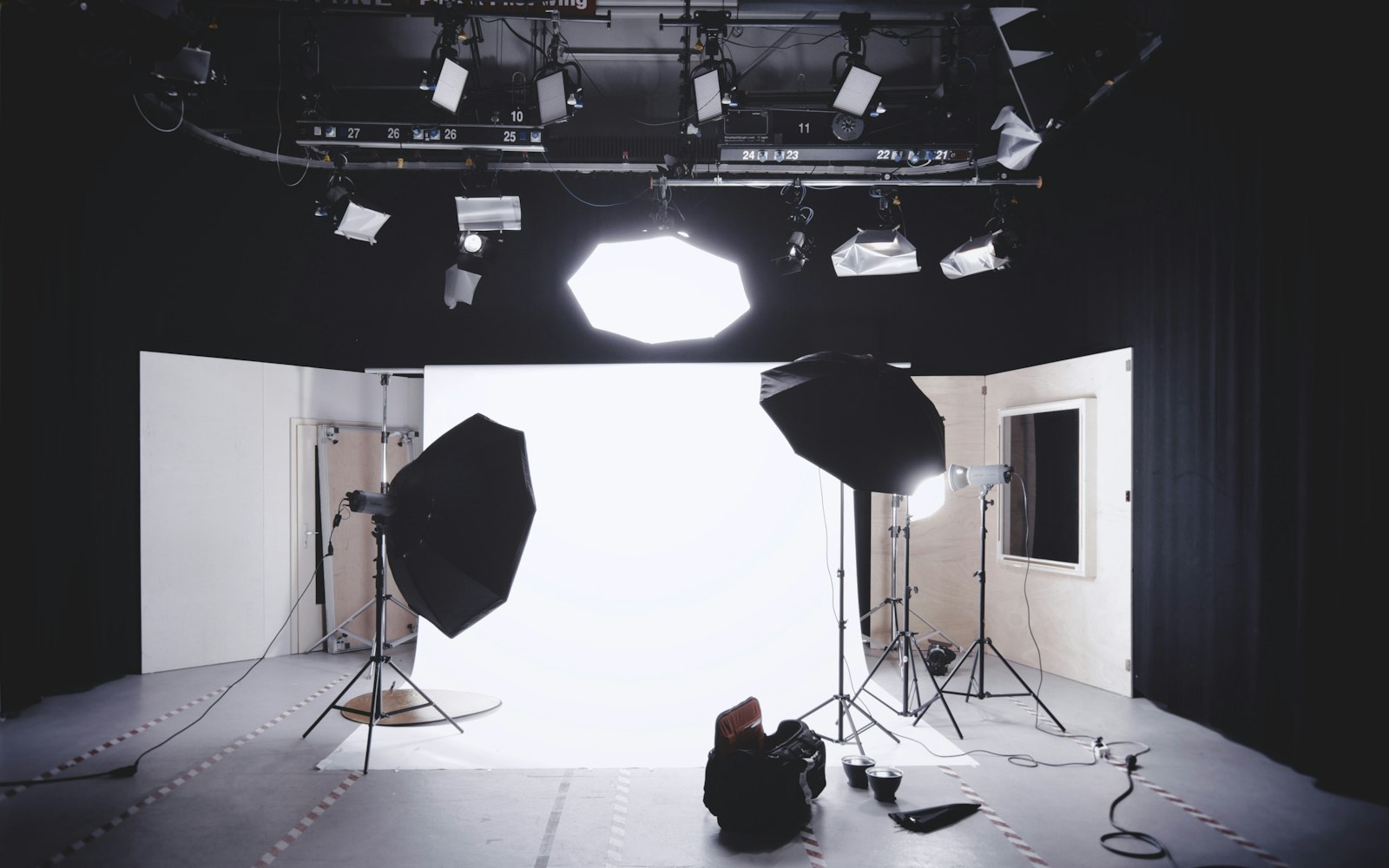 An empty photo studio with a white backdrop, two large octagonal softboxes on stands, an overhead lighting rig and a camera bag on the floor