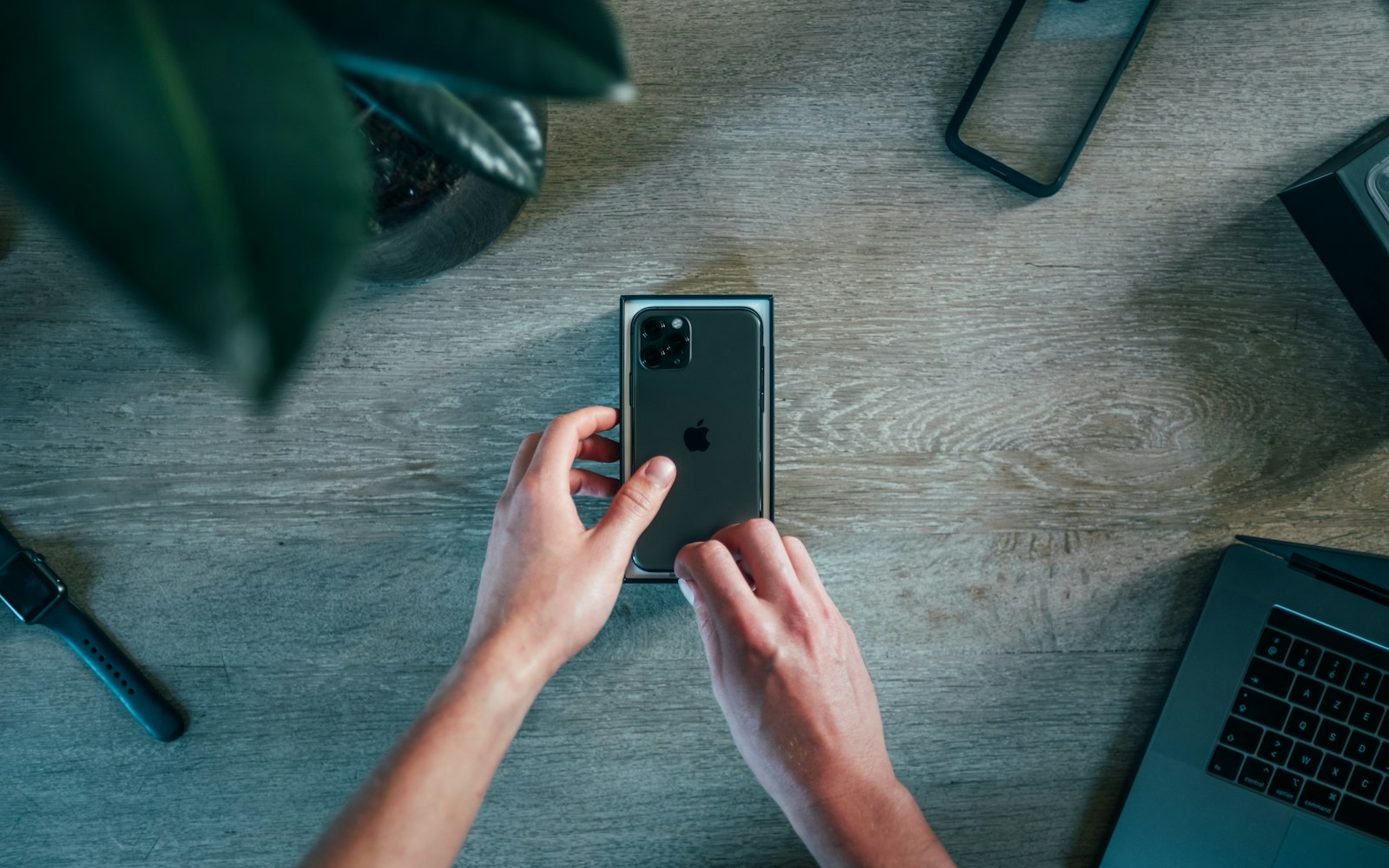 Overhead view of two hands lifting a new smartphone out of its box on a wooden table, beside a plant, a smartwatch and a laptop