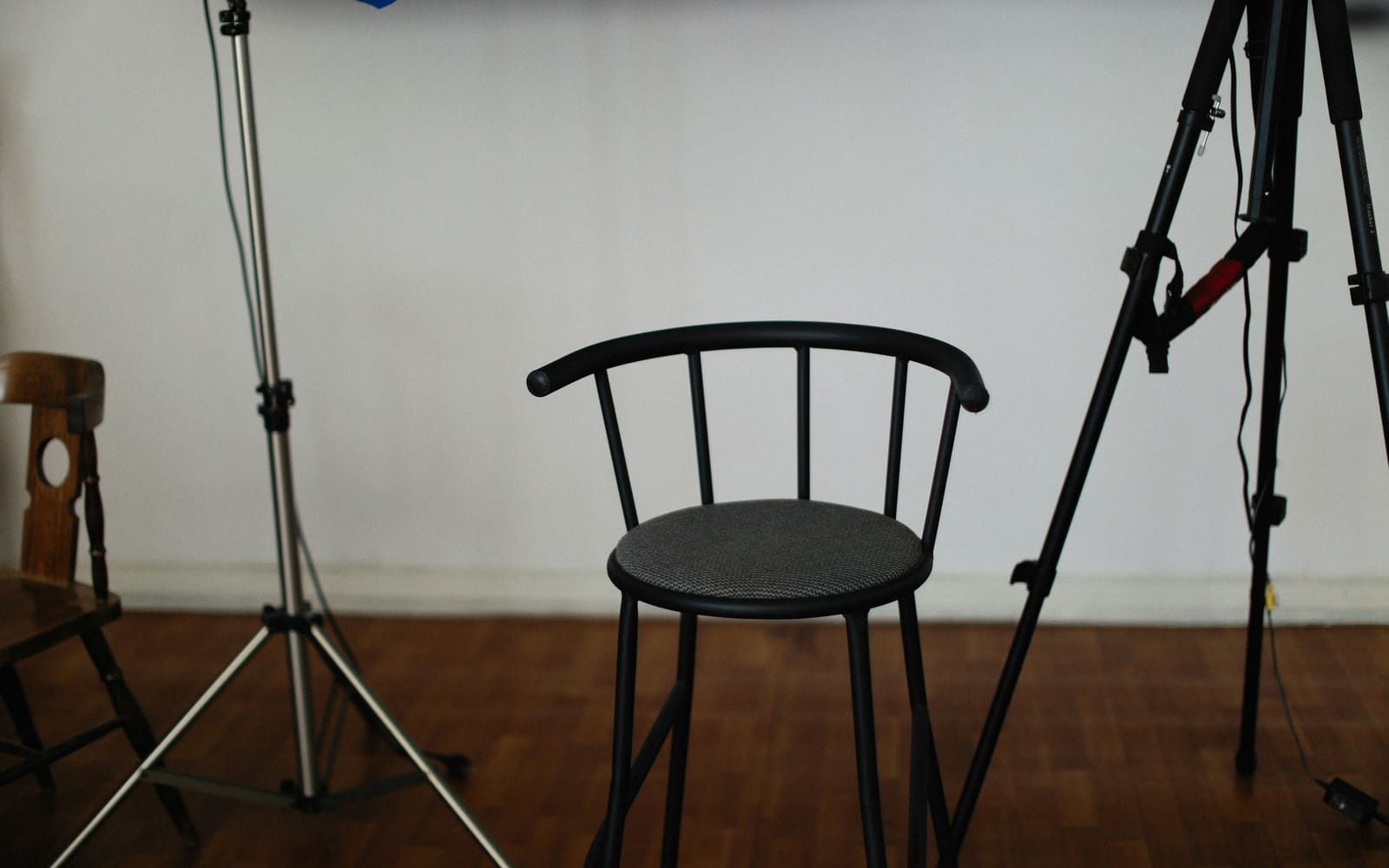 An empty black bar stool stands on a parquet floor in a white-walled room between a silver light stand and a camera tripod