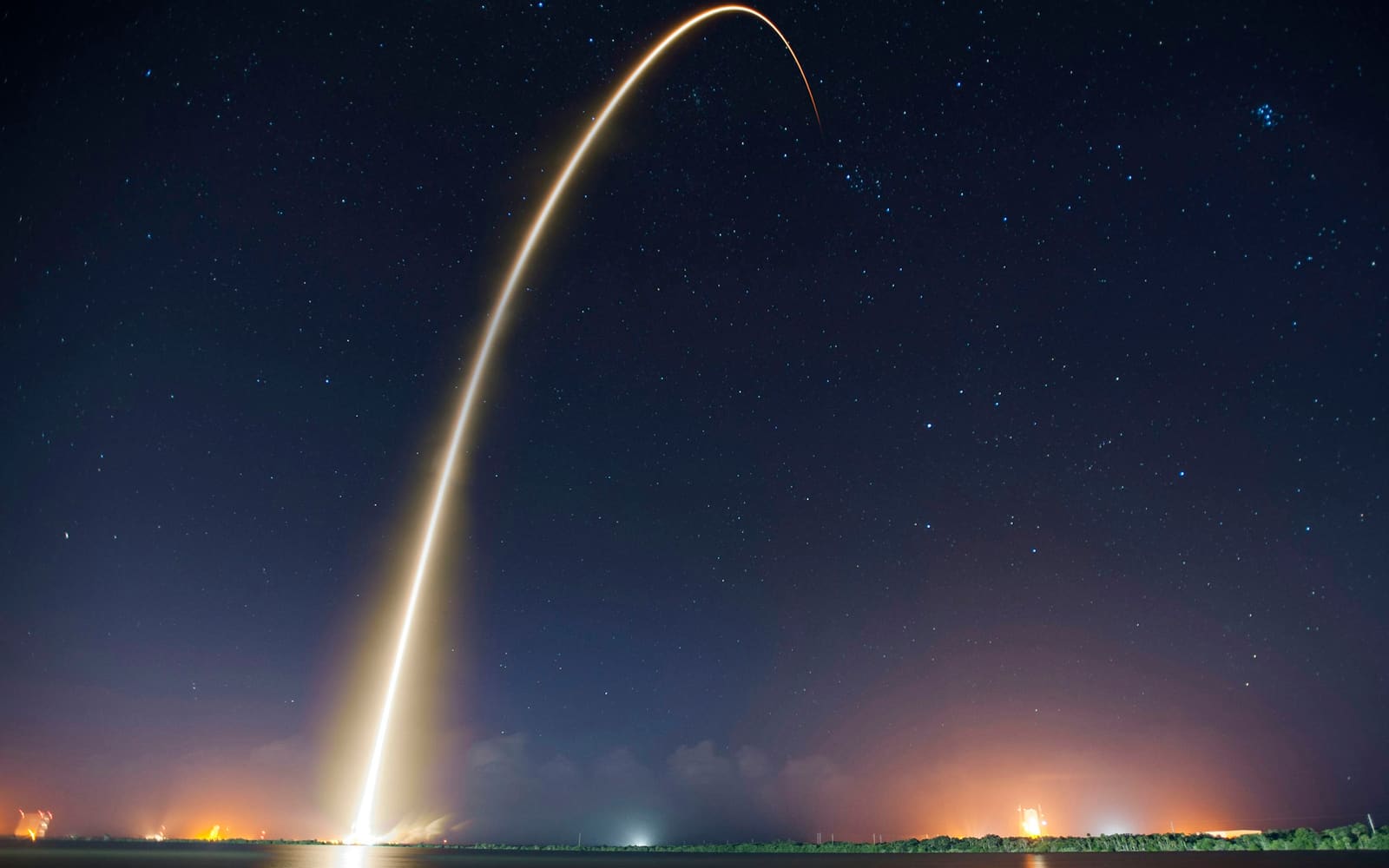 A long-exposure trail of golden light arcs from a distant shoreline into a star-filled night sky, with orange lights glowing low along the water