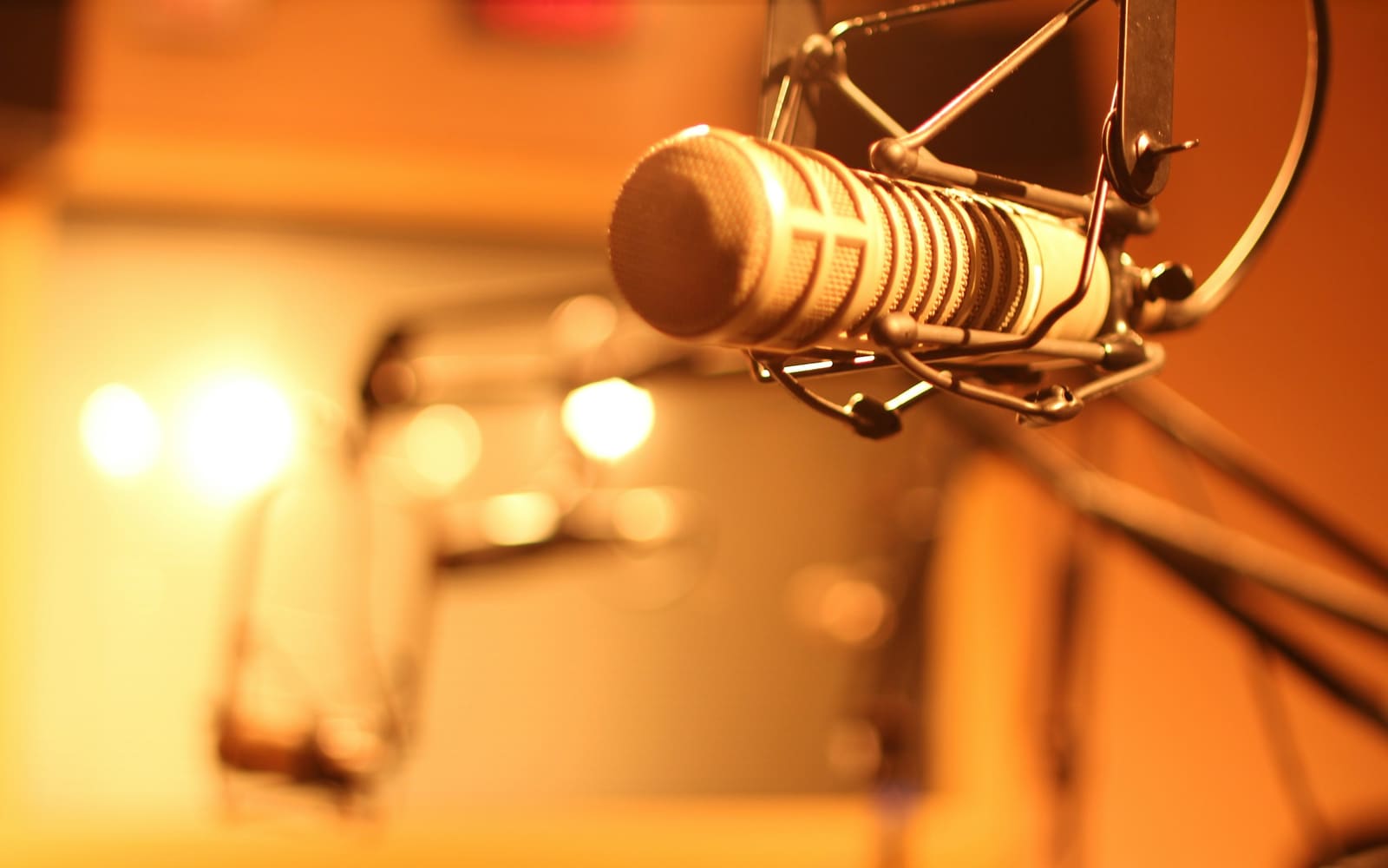 A silver broadcast microphone in a suspension mount on a boom arm, lit in warm amber light with blurred studio lamps glowing behind it