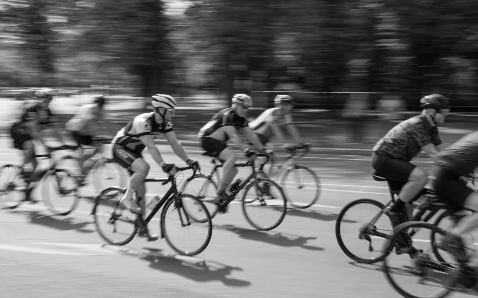 Road cyclists in helmets and jerseys race past in a tight group, shot in black and white with trees blurred by motion behind them