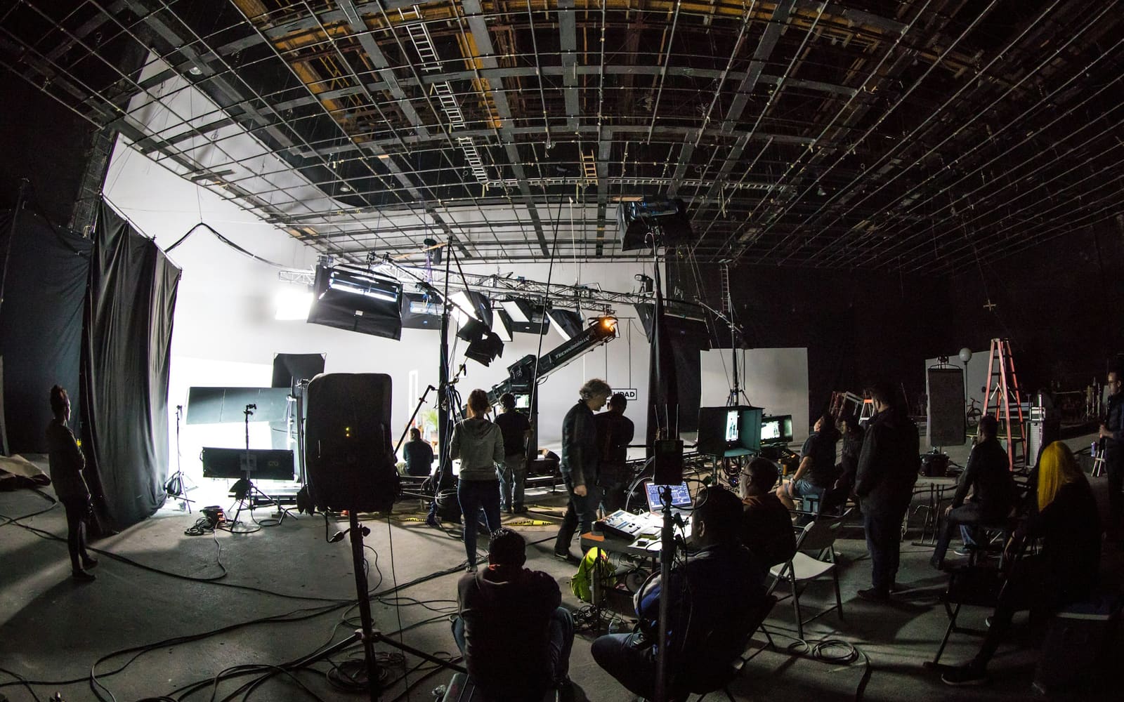 A dim soundstage with a lighting grid overhead, a camera crane, bright studio lights and crew members watching monitors among cables