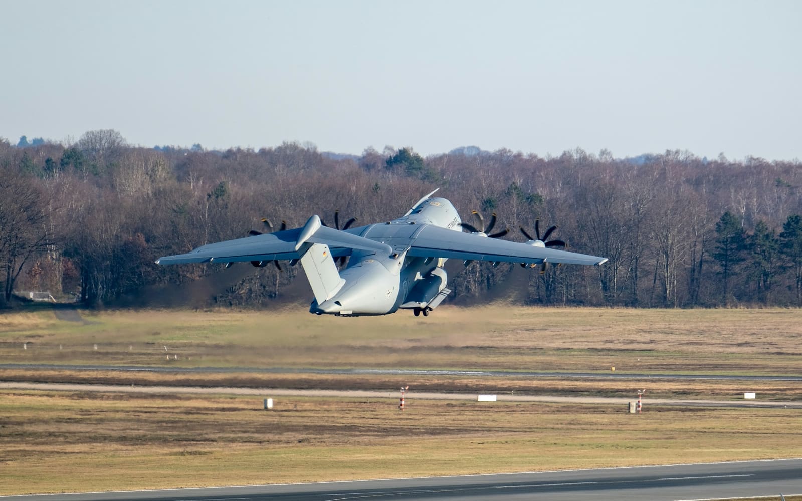 A gray four-engine military transport plane climbs steeply away from a runway, past a brown winter field and a line of bare trees