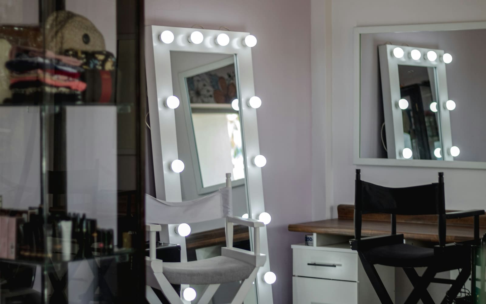 A tall white mirror ringed with glowing round bulbs stands behind a white director’s chair in a pale lilac makeup room, with a second lit mirror on the wall