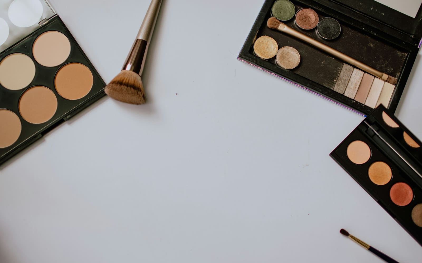 Three open makeup palettes of contour powders and eyeshadows sit at the edges of a pale gray tabletop beside loose makeup brushes