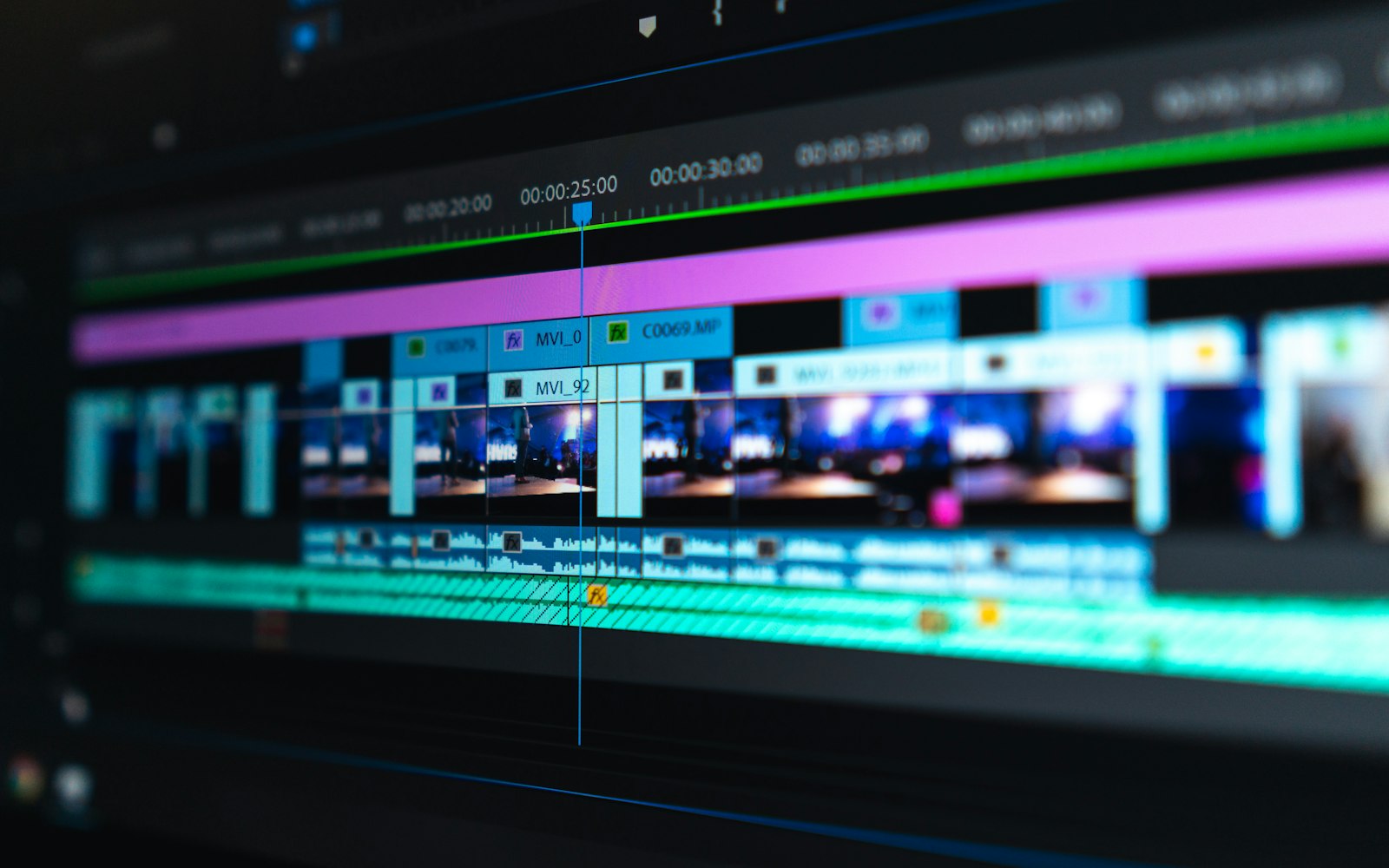 Close-up of a video editing timeline on a dark screen, with a blue playhead crossing stacked purple, blue and green clips and audio tracks