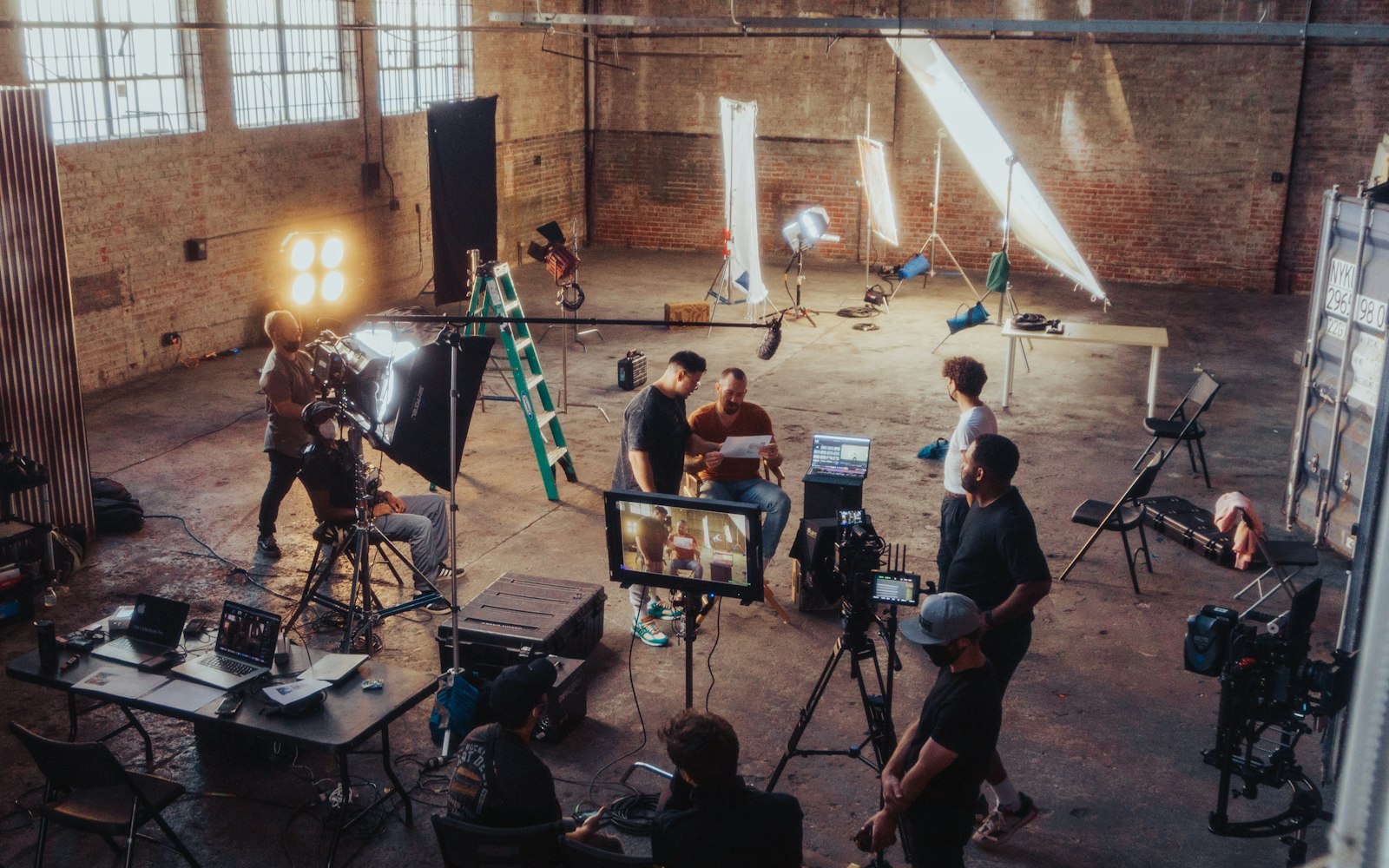 A film crew in a brick warehouse studio with lights, a ladder and cameras, two men reading a script while a monitor shows the shot