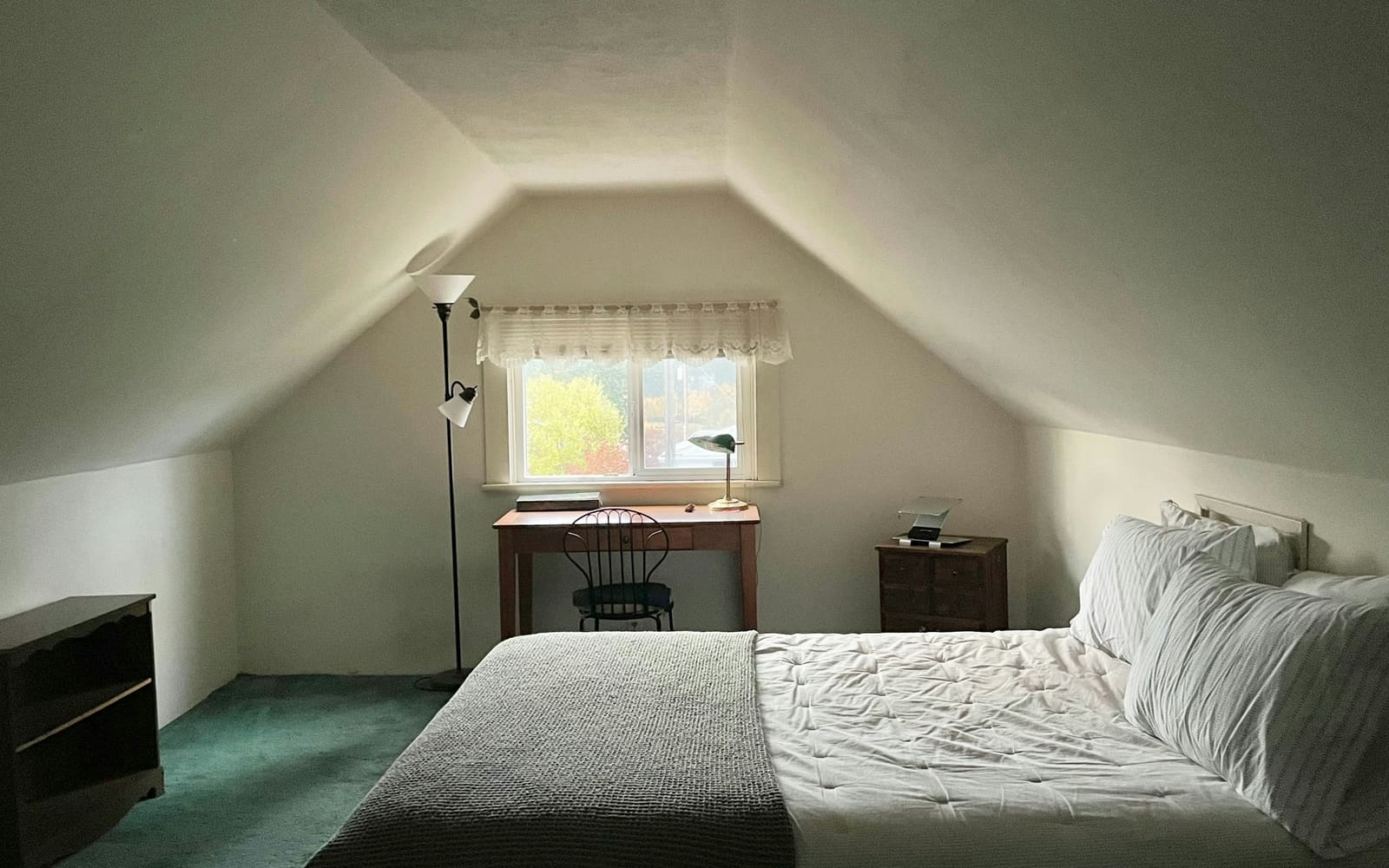 An attic bedroom with sloping white ceilings, a wooden desk and chair beneath a small window, a tall floor lamp and a made bed