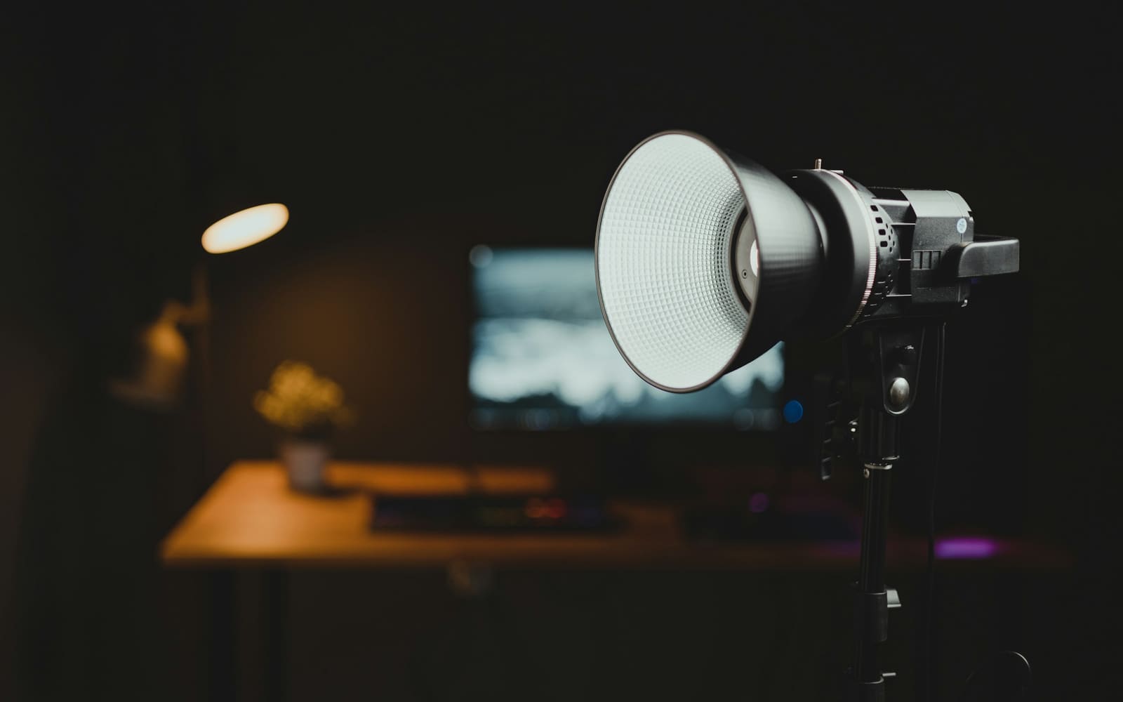 A black studio light with a white reflector dish mounted on a stand in a dark room, a desk, monitor, plant and warm lamp blurred behind it