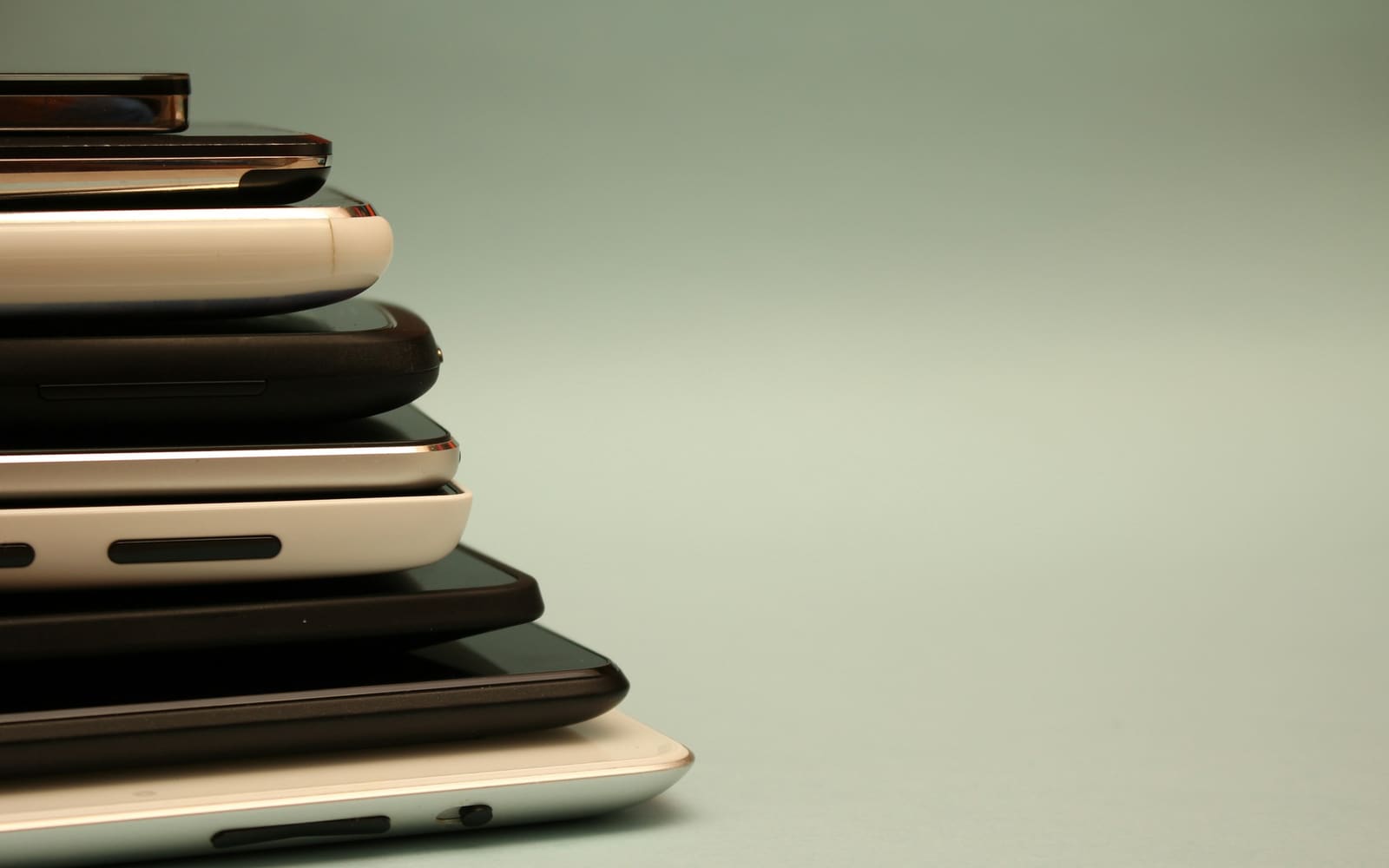 A stack of black, white and silver smartphones and tablets seen from the side, largest at the bottom, against a plain pale green background