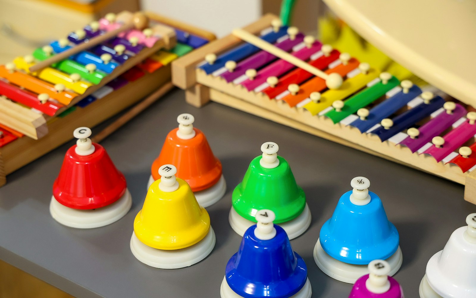 Rainbow-colored desk bells marked with note letters on a gray table, in front of two wooden xylophones with colorful metal bars and mallets
