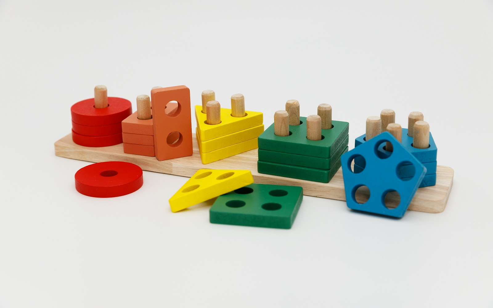 A wooden shape-sorting toy with red, orange, yellow, green and blue stacking shapes on pegs, and a few loose pieces, against a white background
