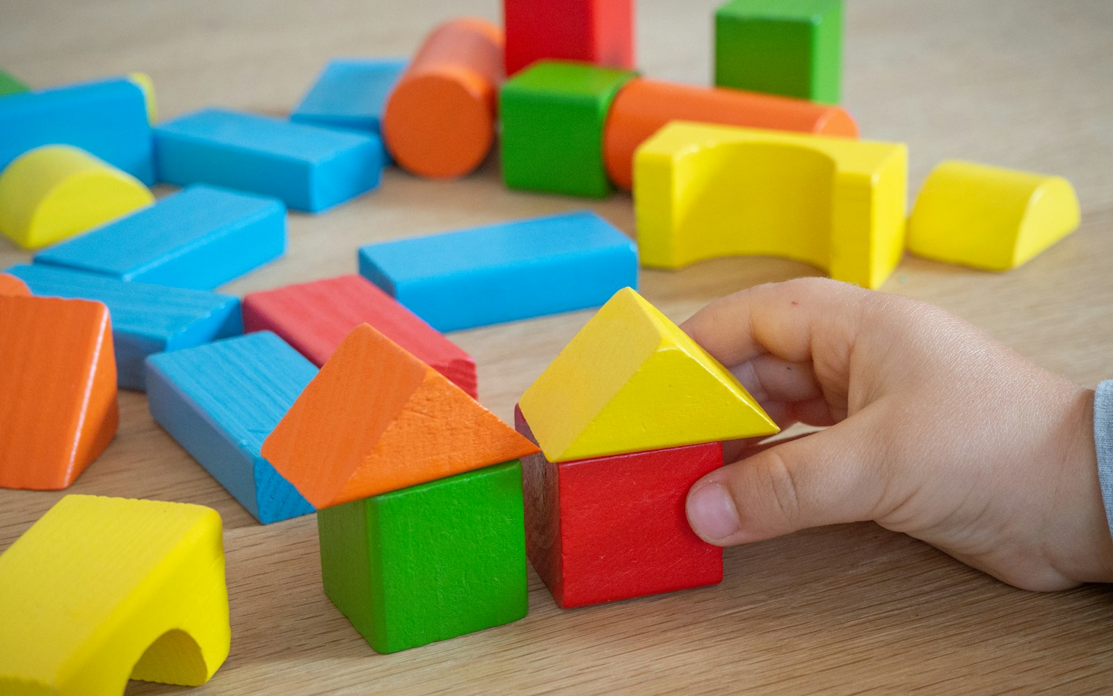 A small child’s hand sets a yellow roof on a red wooden block beside a green-and-orange block house, with more colorful blocks scattered on a table