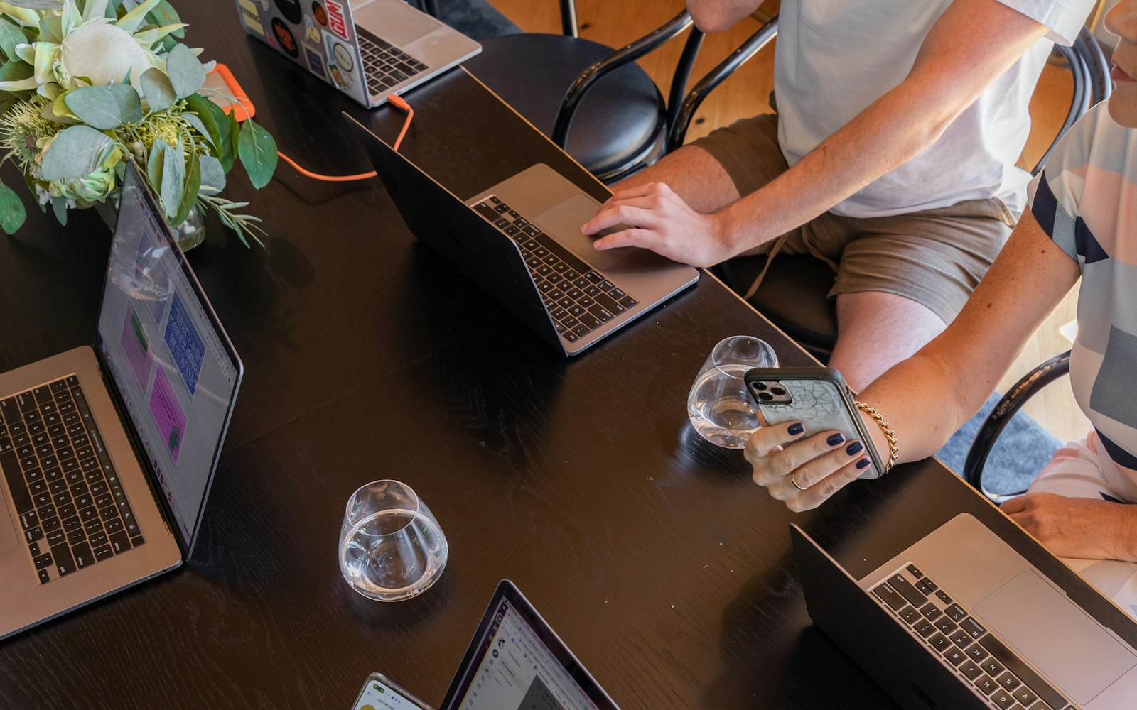 Overhead view of a dark wooden table where several people work on laptops, one holding a smartphone, beside glasses of water and a vase of pale flowers and eucalyptus