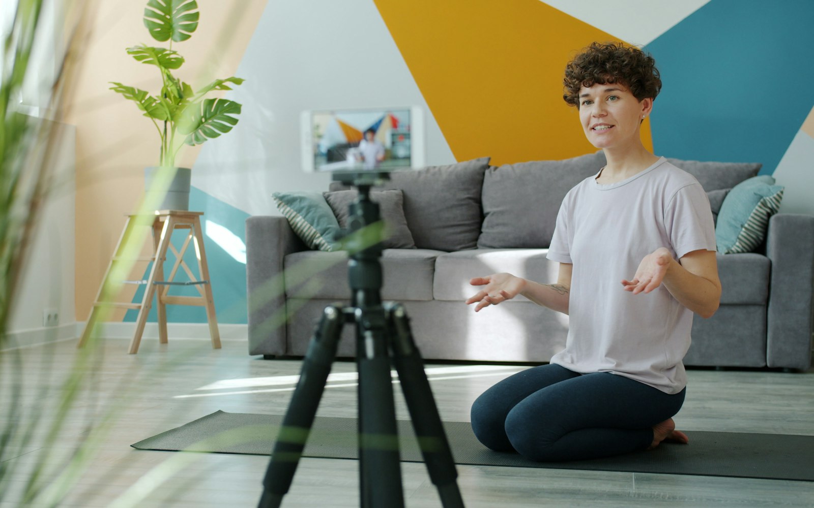 A woman kneeling on a yoga mat in a brightly painted living room talks to a smartphone on a tripod, which shows her on its screen