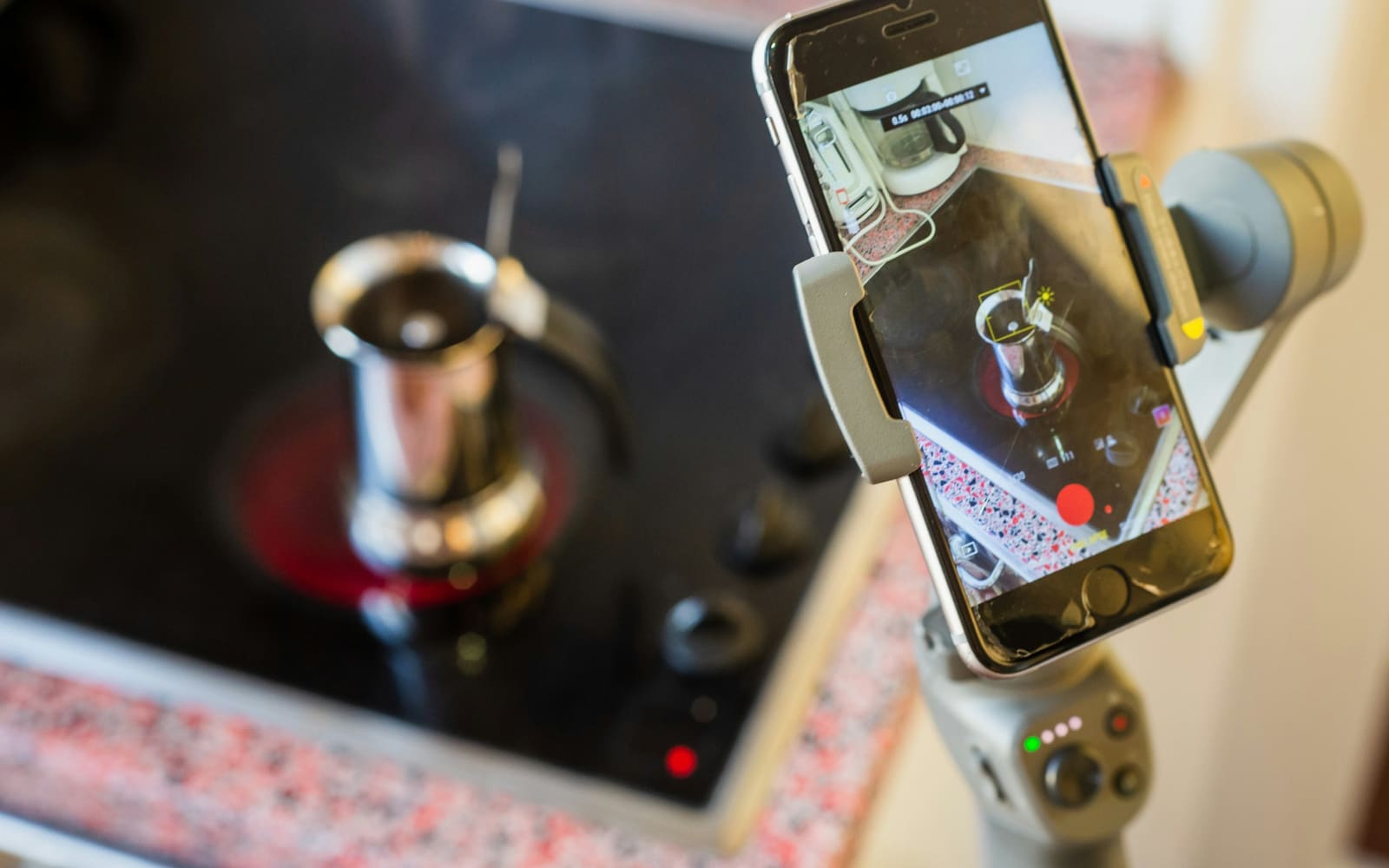 A smartphone on a gray handheld gimbal records a steel coffee pot heating on a black ceramic stovetop, the phone screen showing the shot