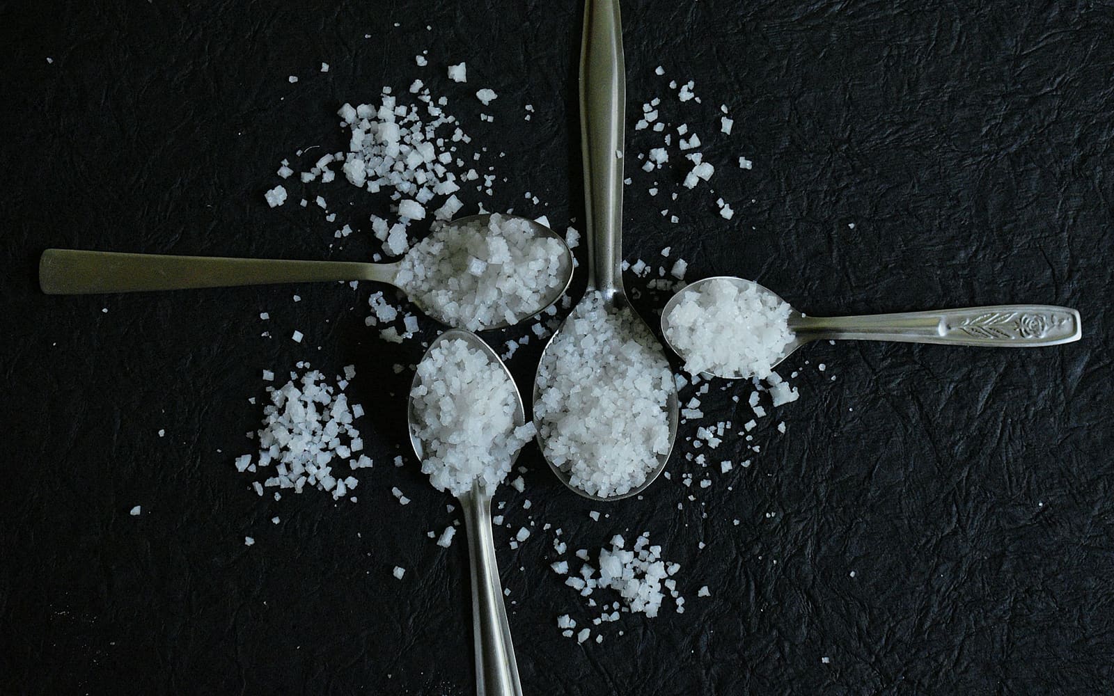Four metal spoons heaped with coarse white salt crystals are arranged in a cross on a dark crinkled surface, with loose crystals scattered around them