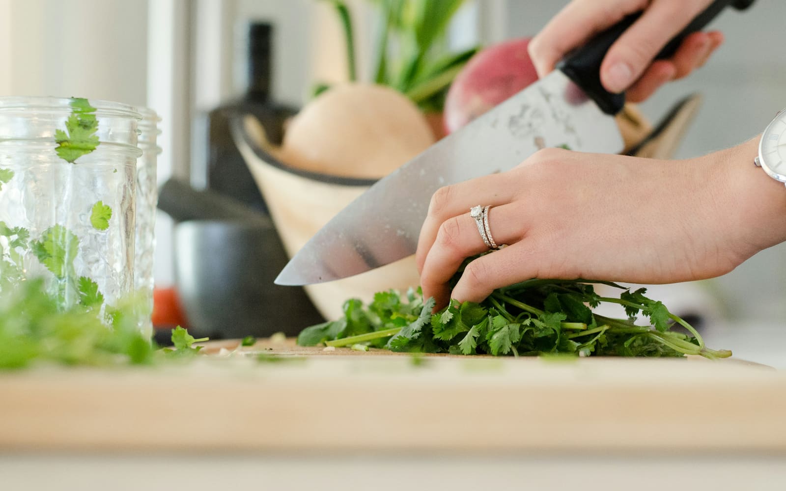 Hands chop a bunch of fresh cilantro with a large chef’s knife on a wooden cutting board, with a glass jar and a bowl of vegetables behind